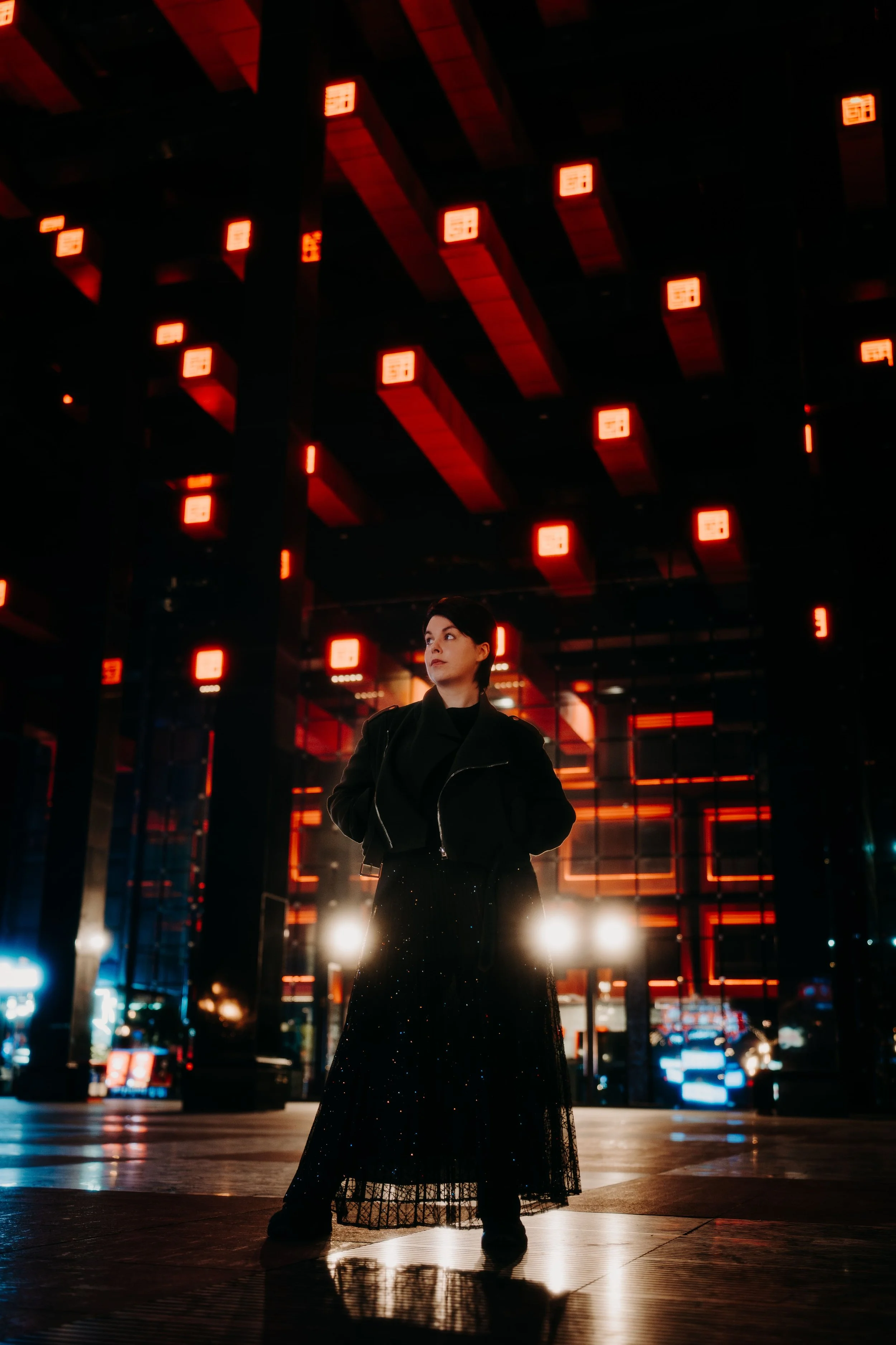 Une femme debout dans une zone urbaine nocturne avec des lumières rouges en arrière-plan, portant une veste noire et une jupe longue en tulle noir, sous un plafond éclairé aux néons rouges.