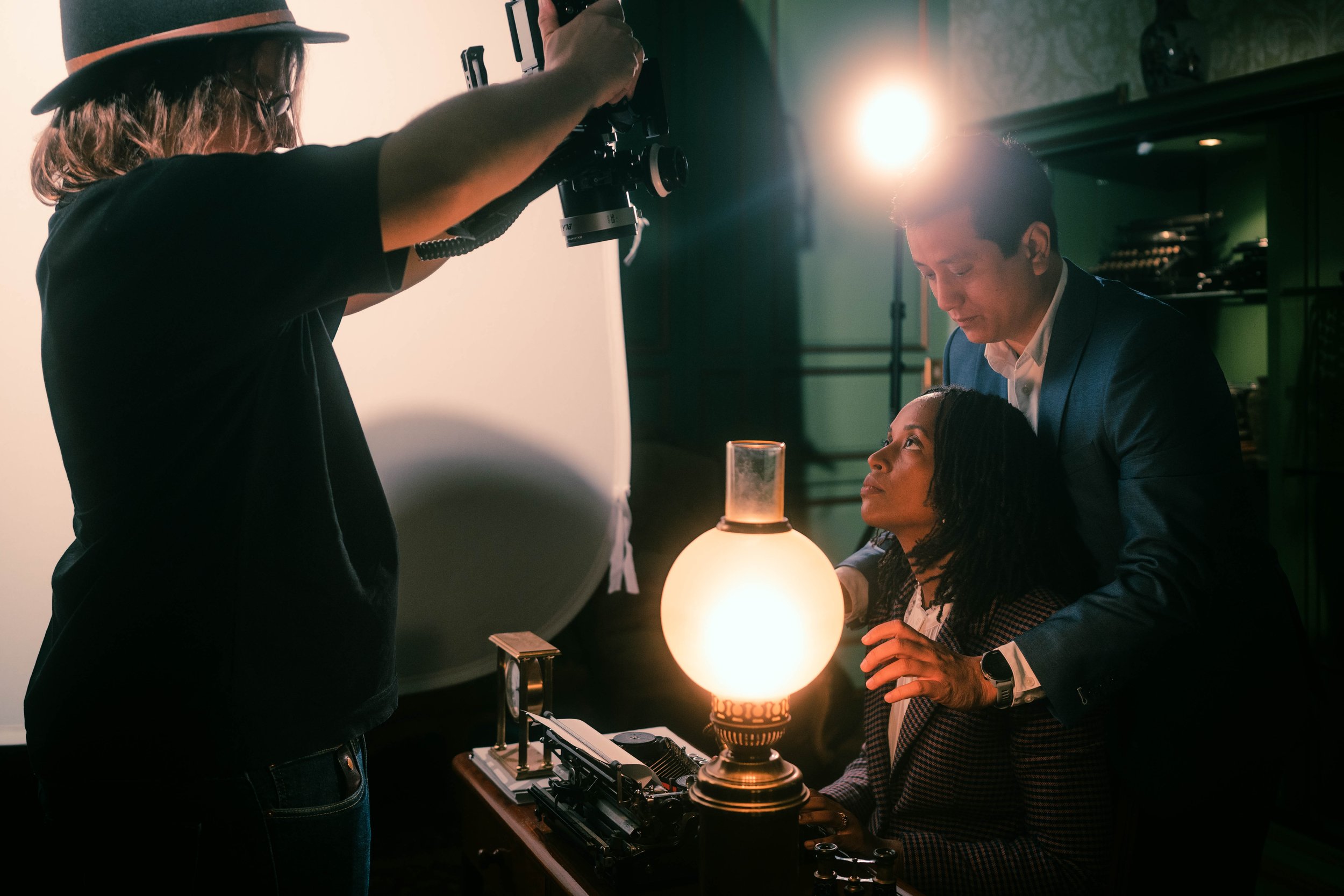 Une séance de photographie en studio avec un photographe, une femme assise, un homme debout, et un assistant tenant un appareil photo, illuminée par une lampe