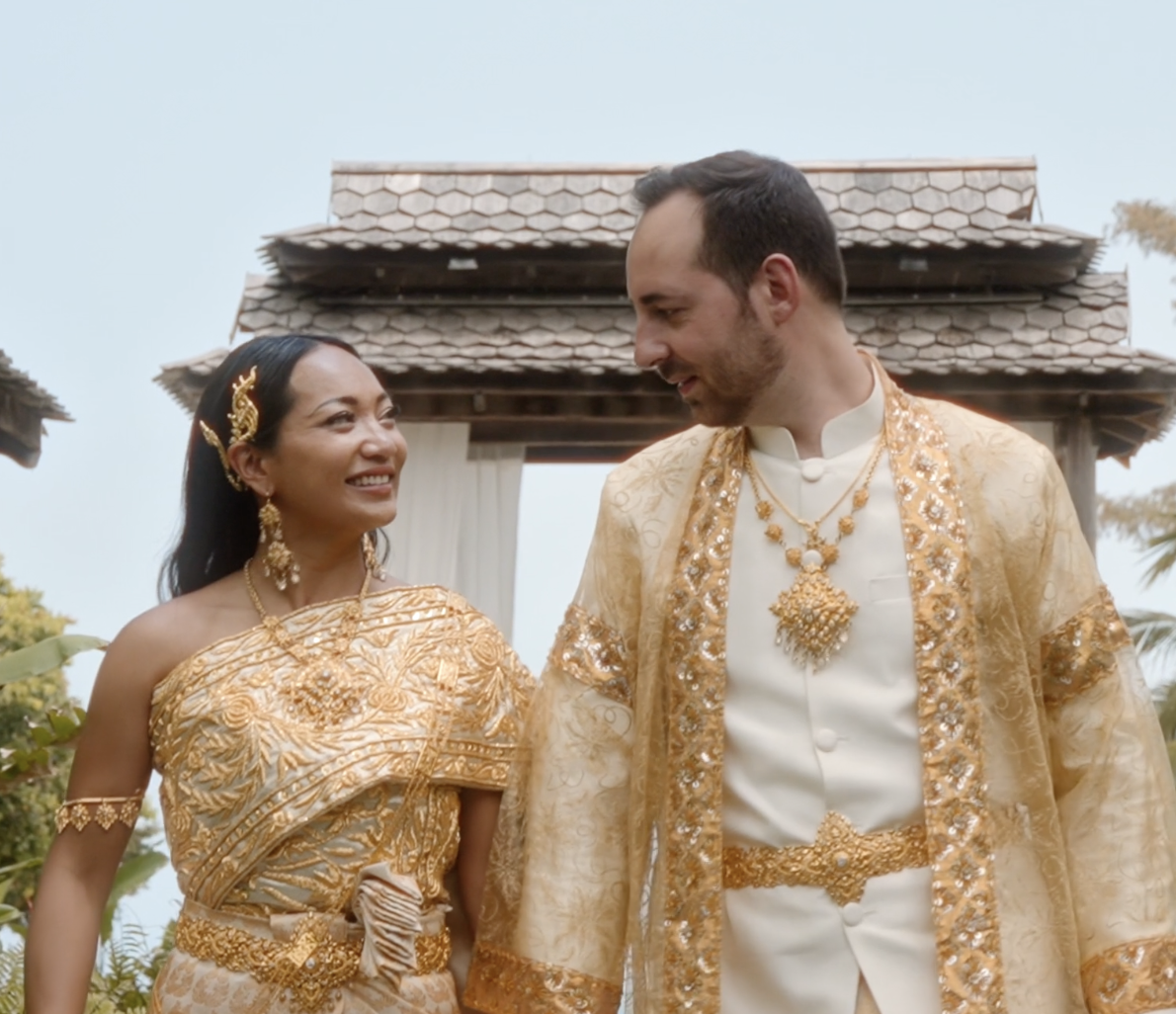 Un couple en vêtements traditionnels dorés lors d'un mariage, se regardant avec sourire devant une structure en bois, dans un cadre extérieur