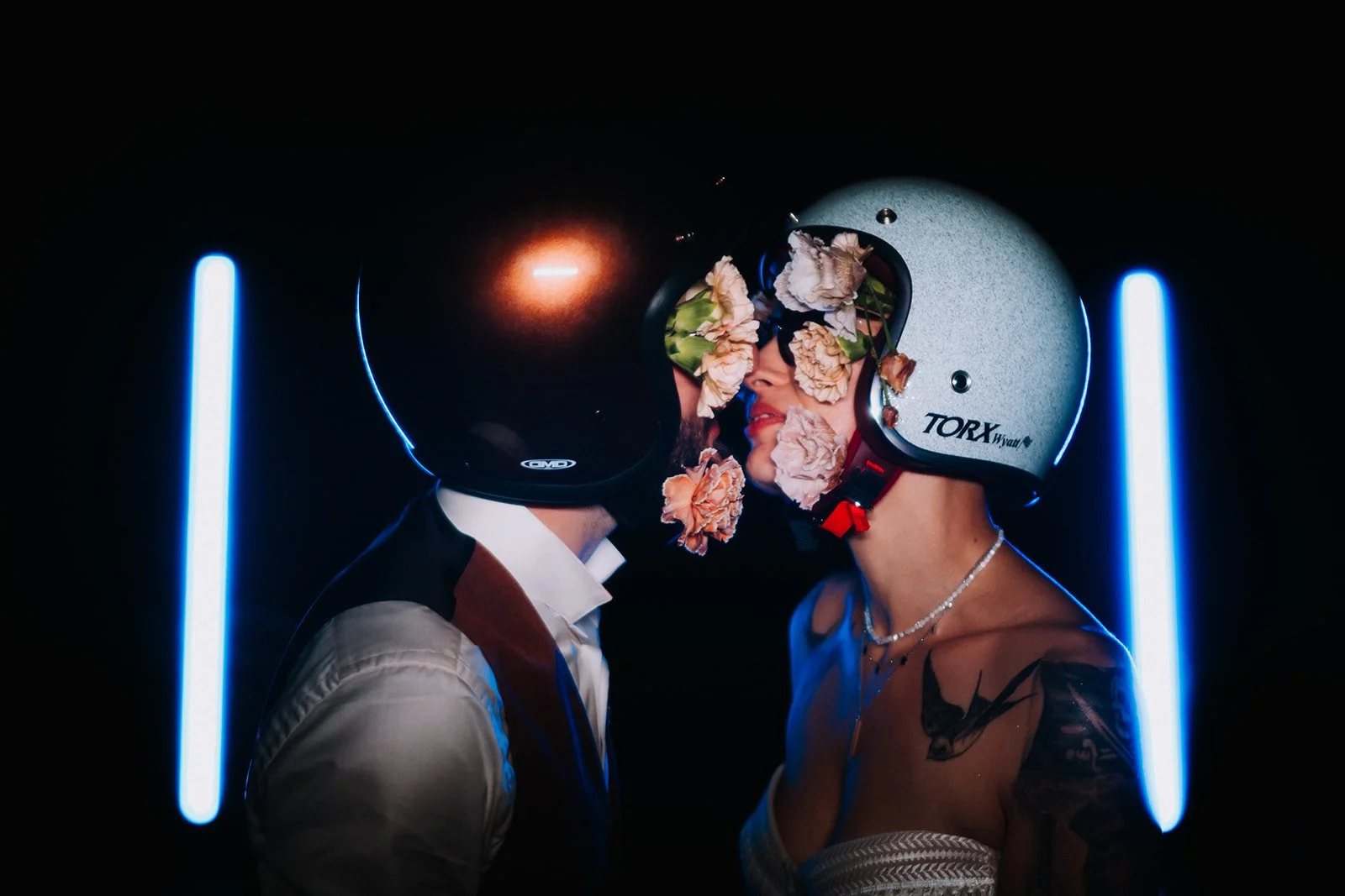 Un couple portant des casques de motos, face à face, le visage de la femme décoré de fleurs, l'ambiance est sombre avec deux lumières bleues à l'arrière.