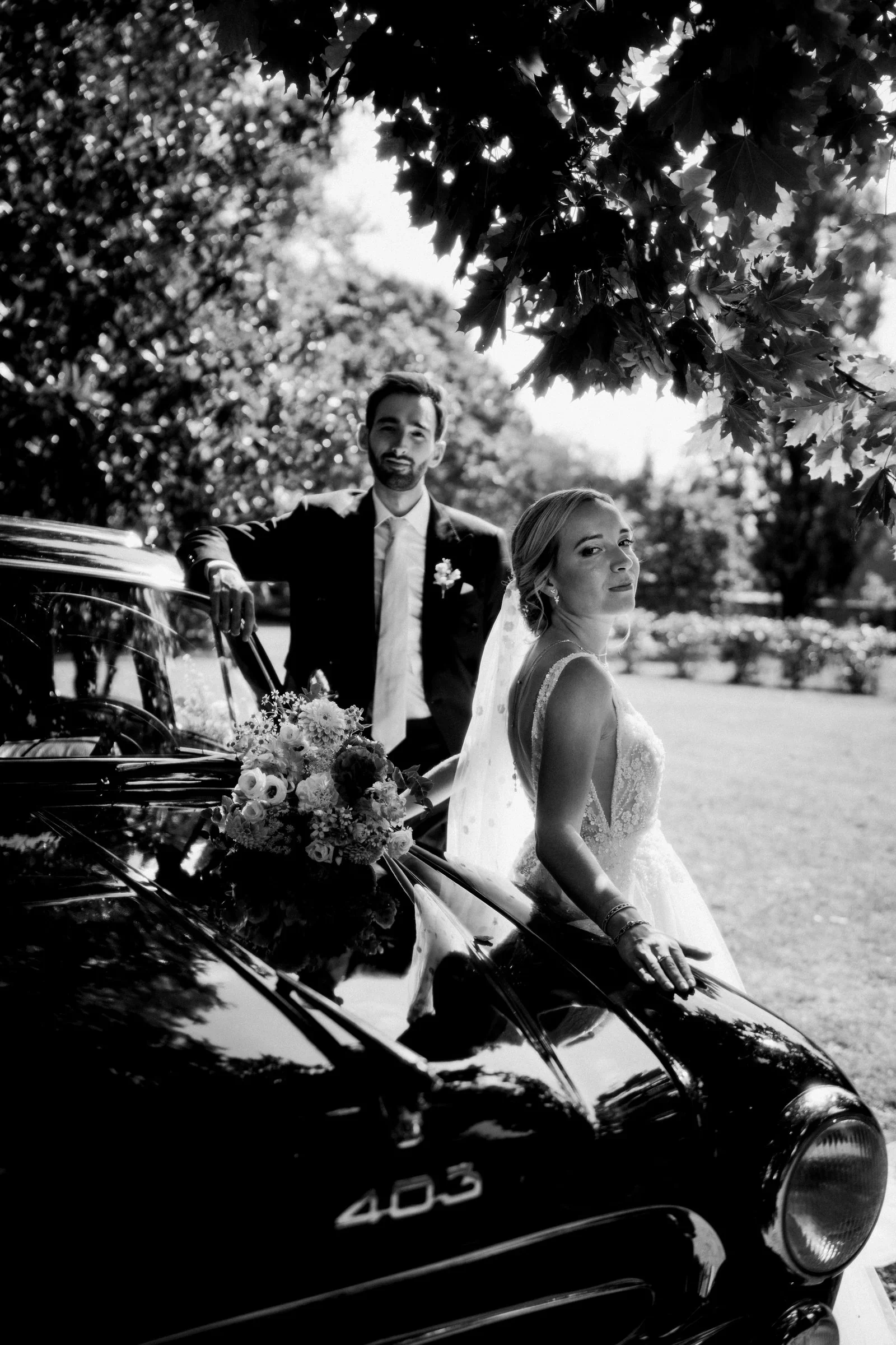couple-maries-voiture-ancienne-peugeot-403-jardin-noir-blanc-photographe-mariage-film-mariage-the-wild-strawberry.webp