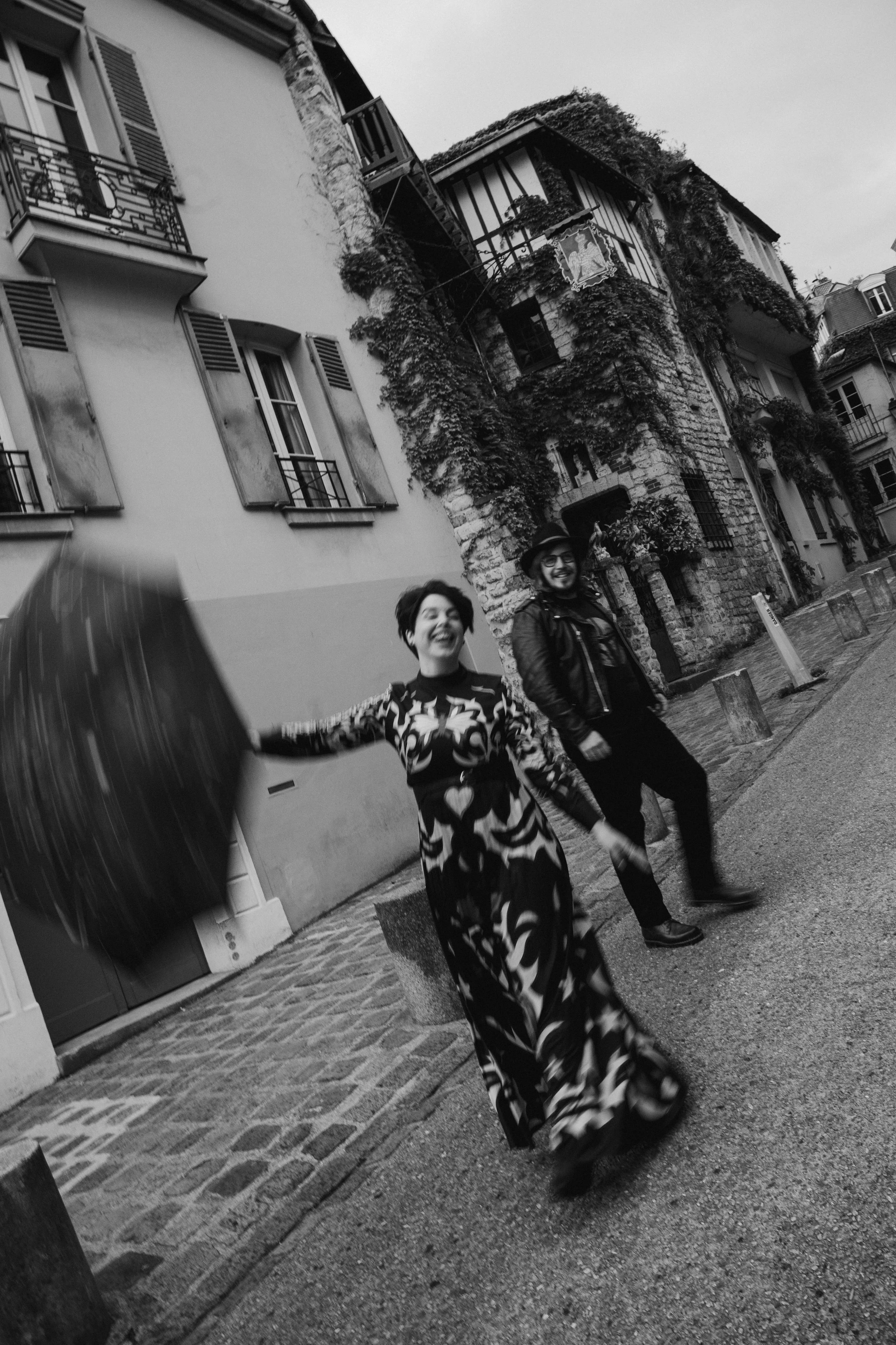 Deux personnes souriantes marchant dans une rue pavée, avec des bâtiments anciens couverts de lierre en arrière-plan, sous un ciel nuageux, en noir et blanc.