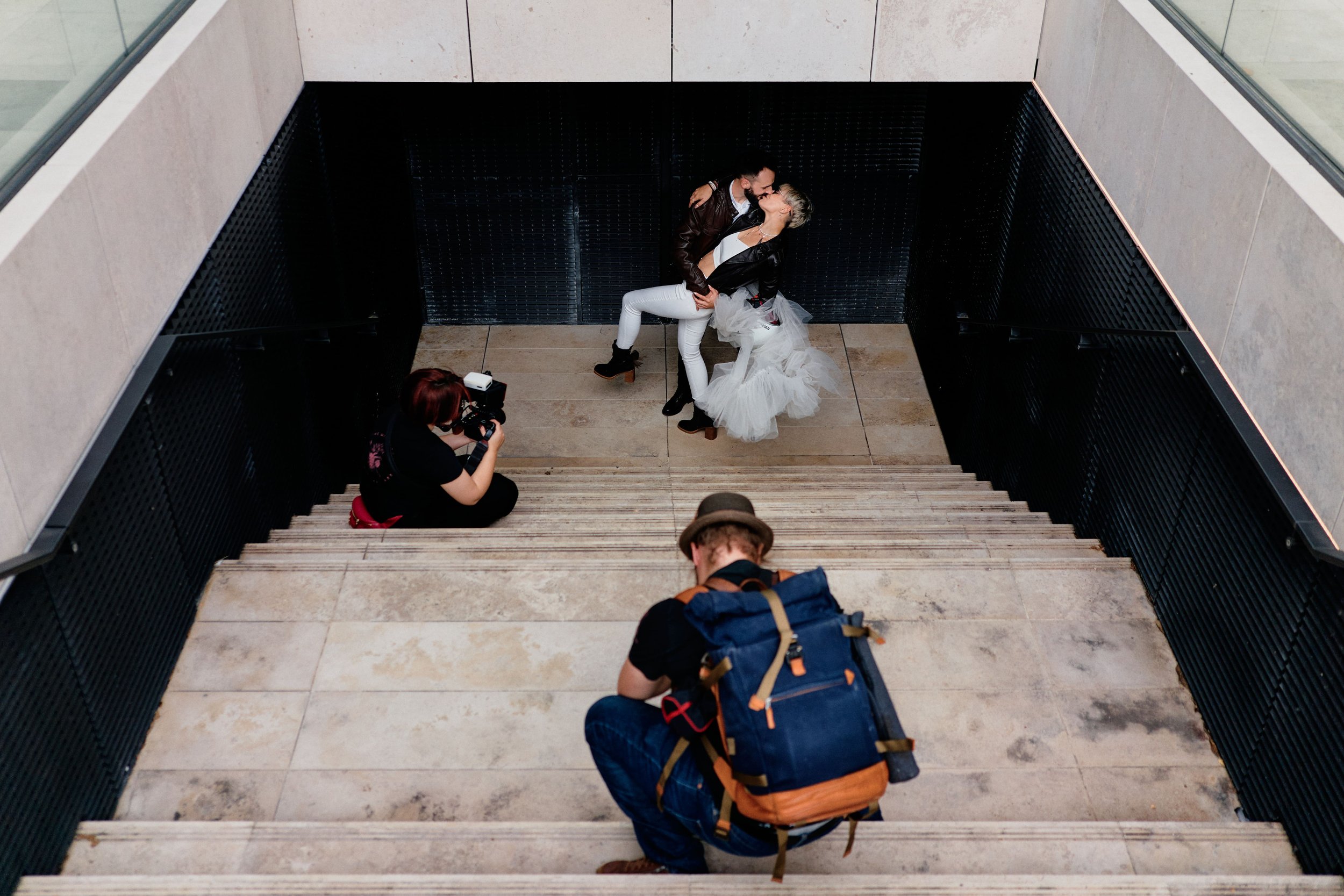 Deux personnes en mariage posent pour une photo, une femme en robe de mariée blanche et un homme en veste en cuir, dans un espace intérieur. Un photographe est accroupi avec un appareil photo, et un autre homme avec un sac à dos regarde la scène depu