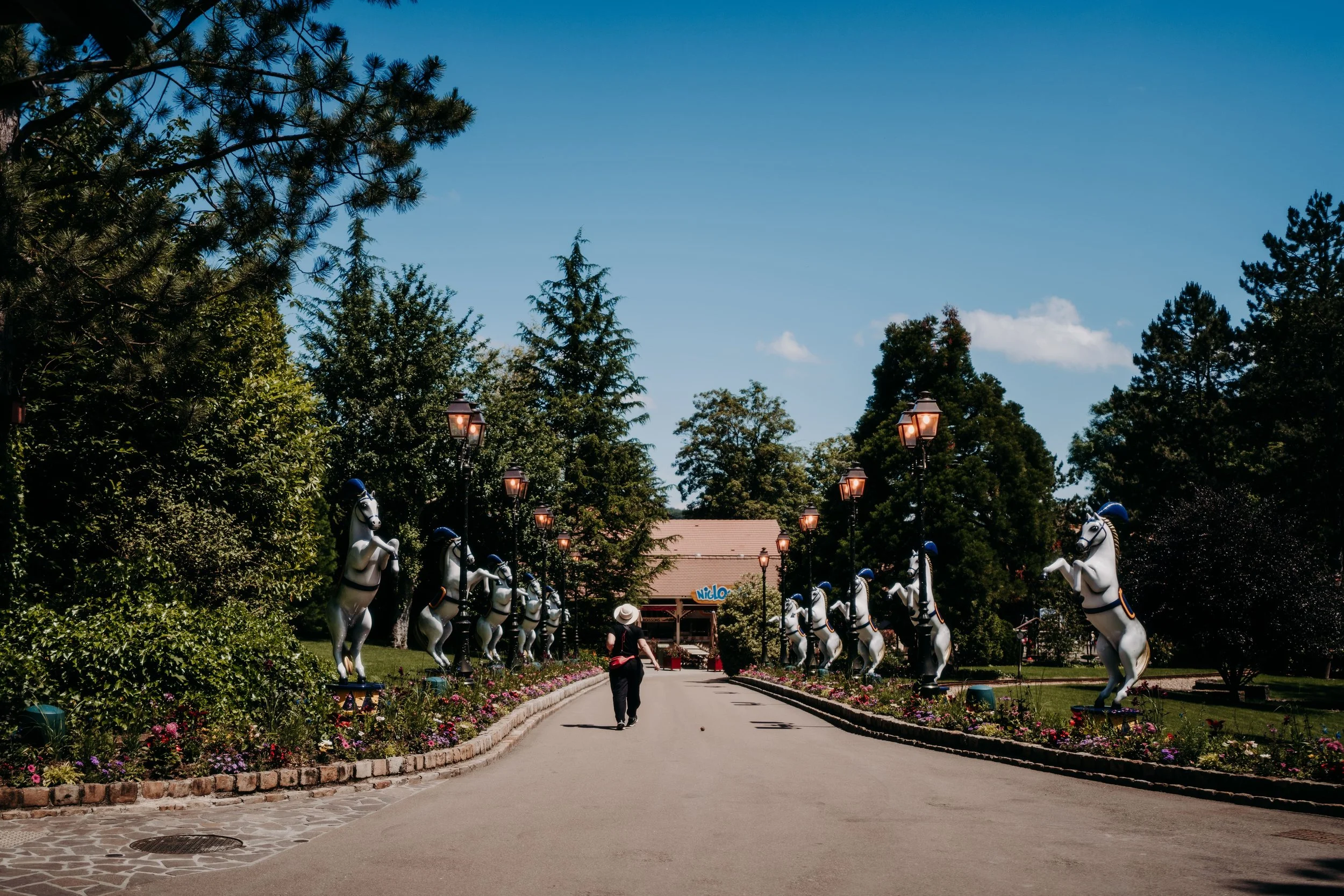 Allée bordée de statues de chevaux en plastique avec lampadaires, arbres et un bâtiment en arrière-plan, sous un ciel bleu.