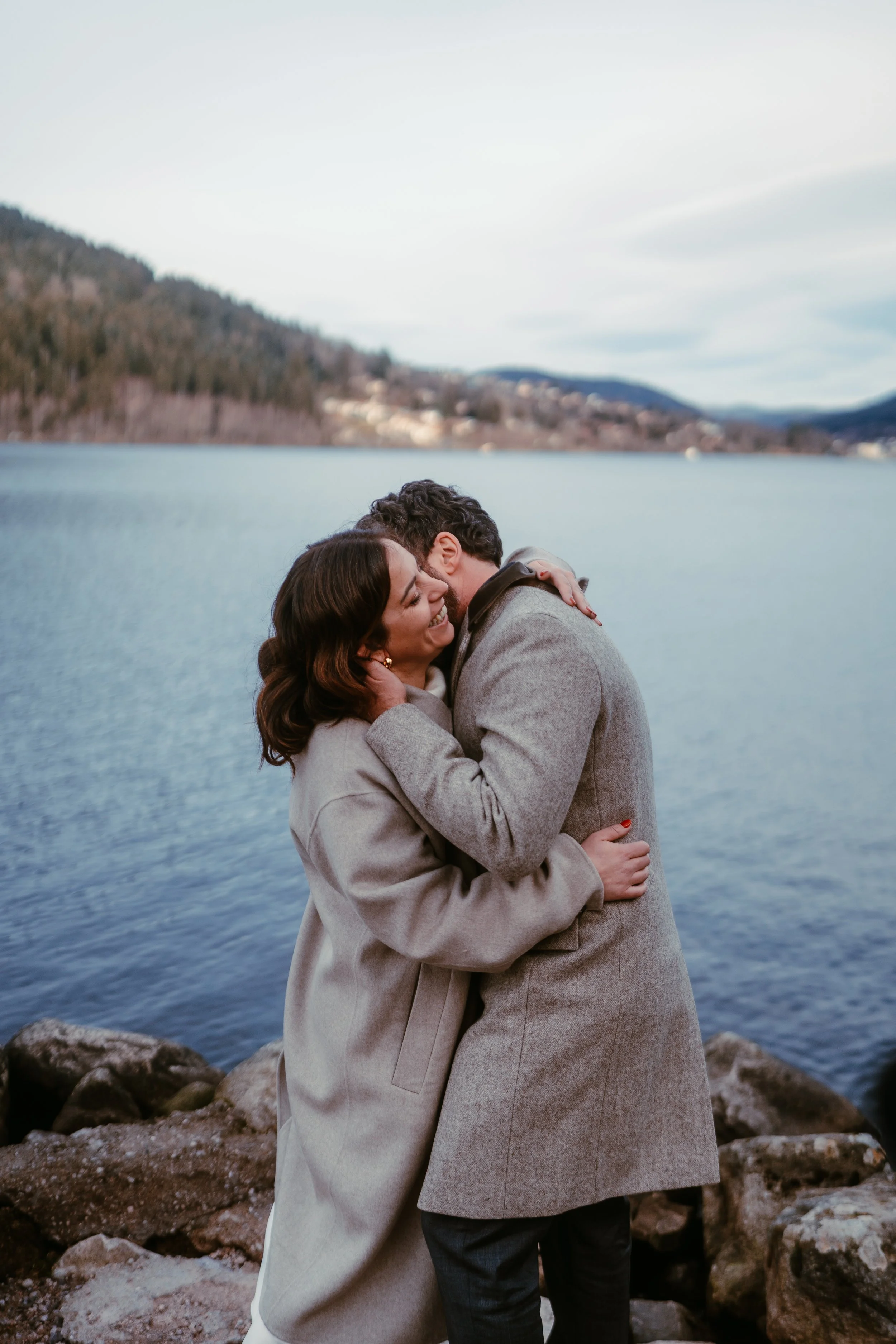 Un couple s'embrasse près d'un lac, en hiver avec des montagnes en arrière-plan.