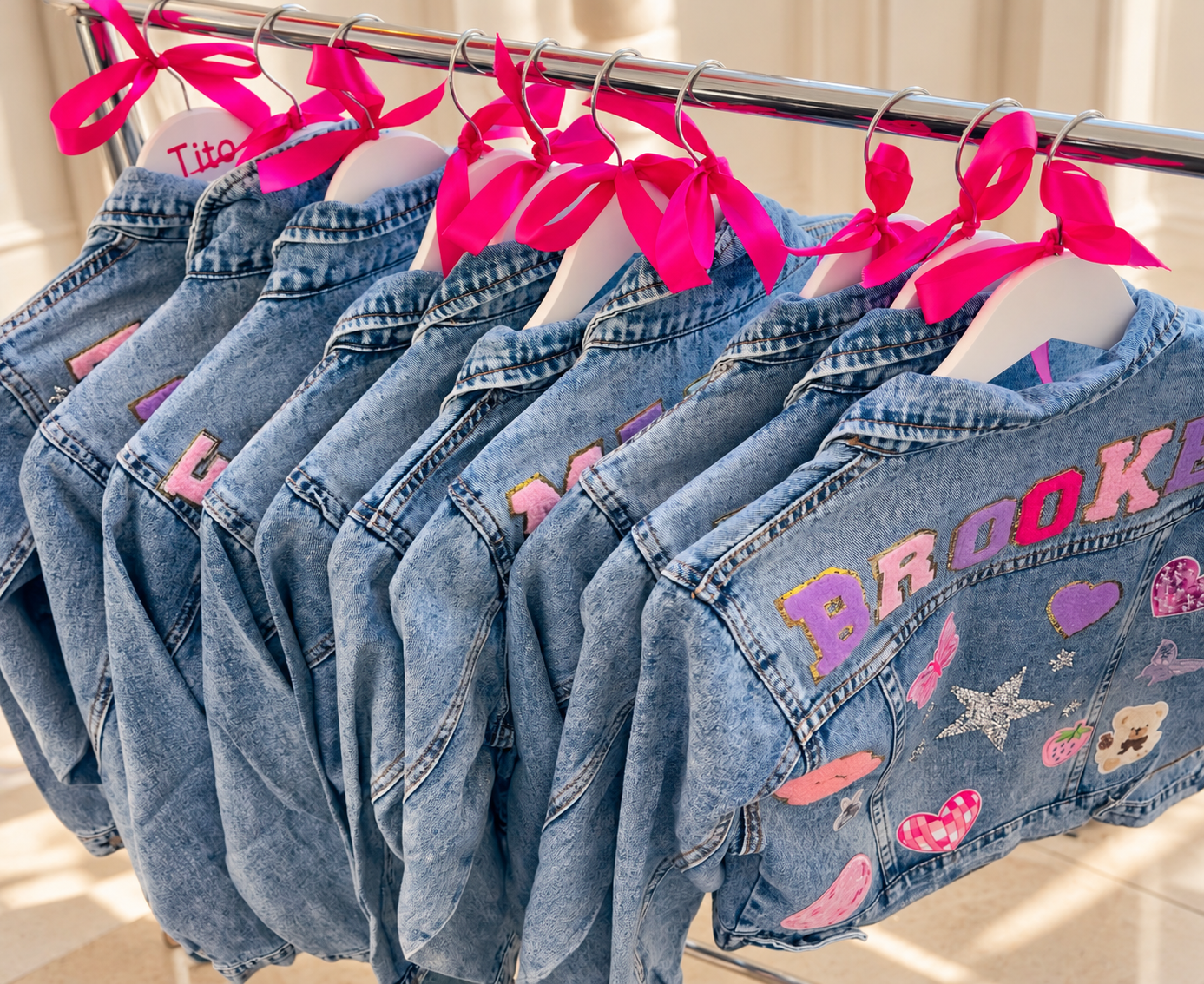 Personalised denim jackets with colourful name and motif patches, displayed on white hangers with bright pink bows at a Luvlybubbly patch party.