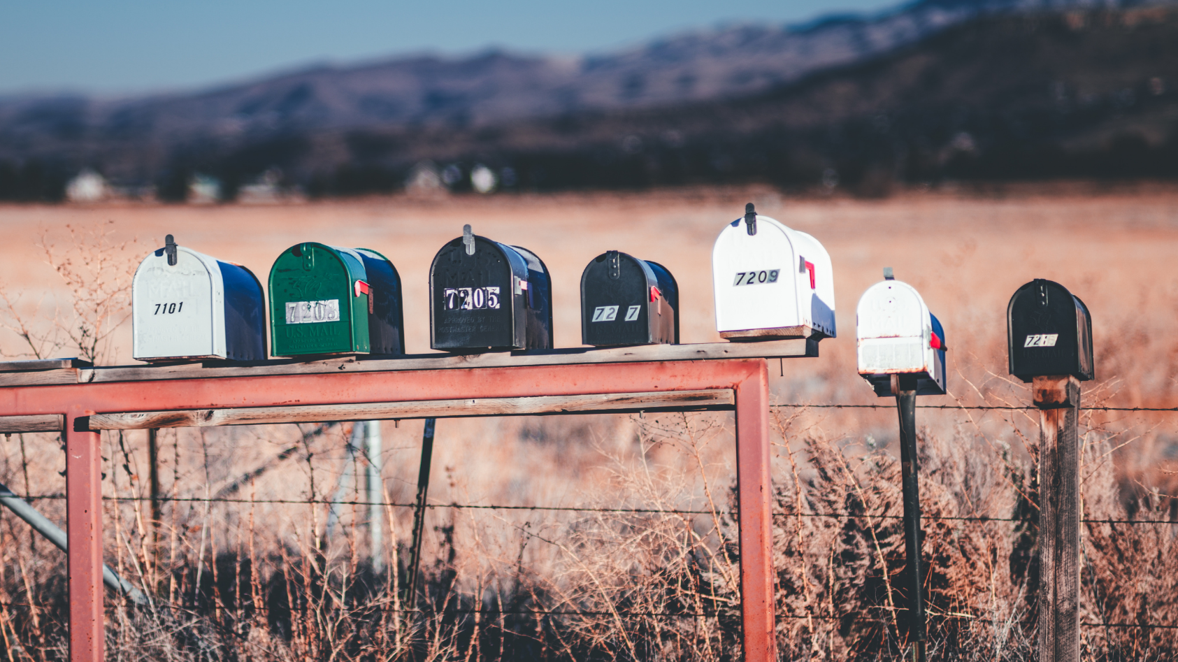A row of eight mailboxes on a wooden platform outdoors in a rural area with dry grass and mountains in the background.