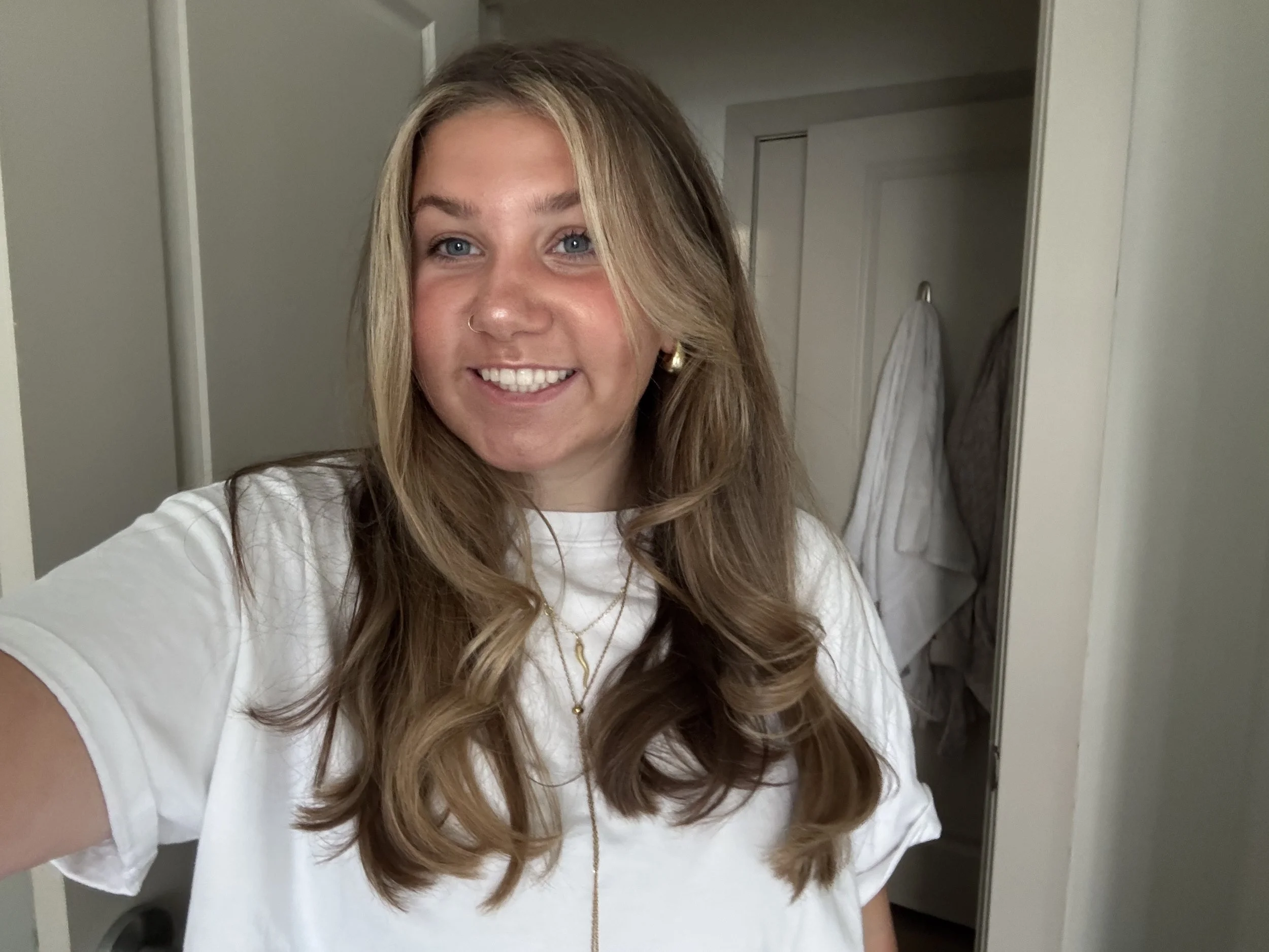 A selfie of a young woman in a white shirt with long dirty blonde hair, smiling indoors