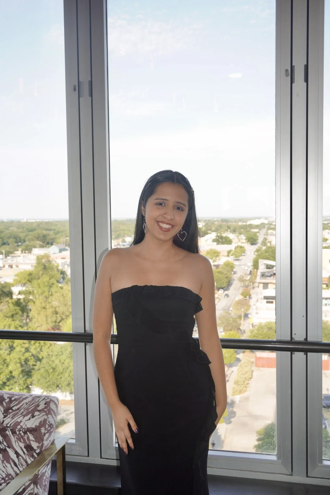 A young woman with long brown hair, smiling infront of some windows, in a strapless black dress