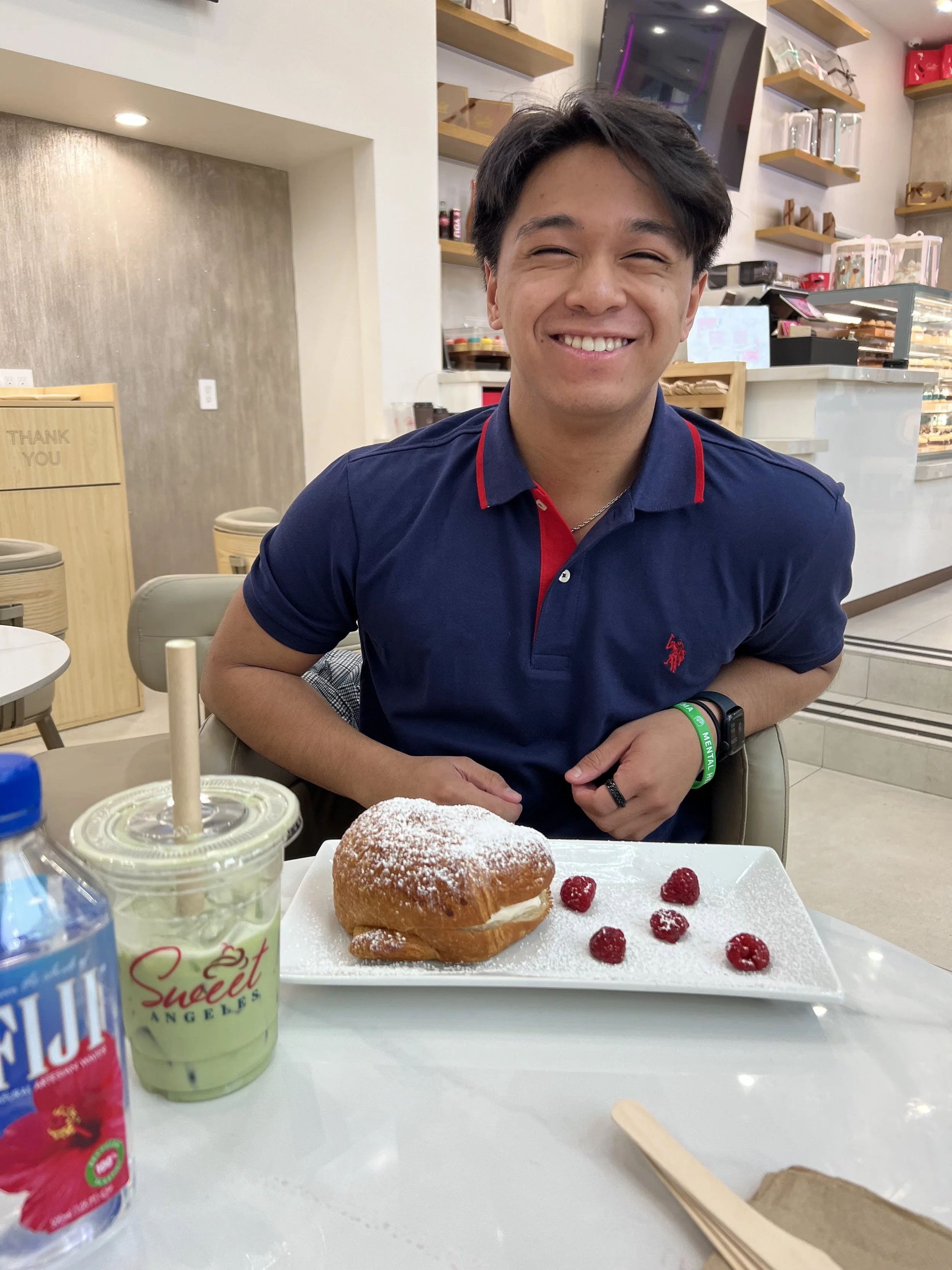 A young man sitting at a table indoors, smiling infront of some food and drinks, with short dark hair