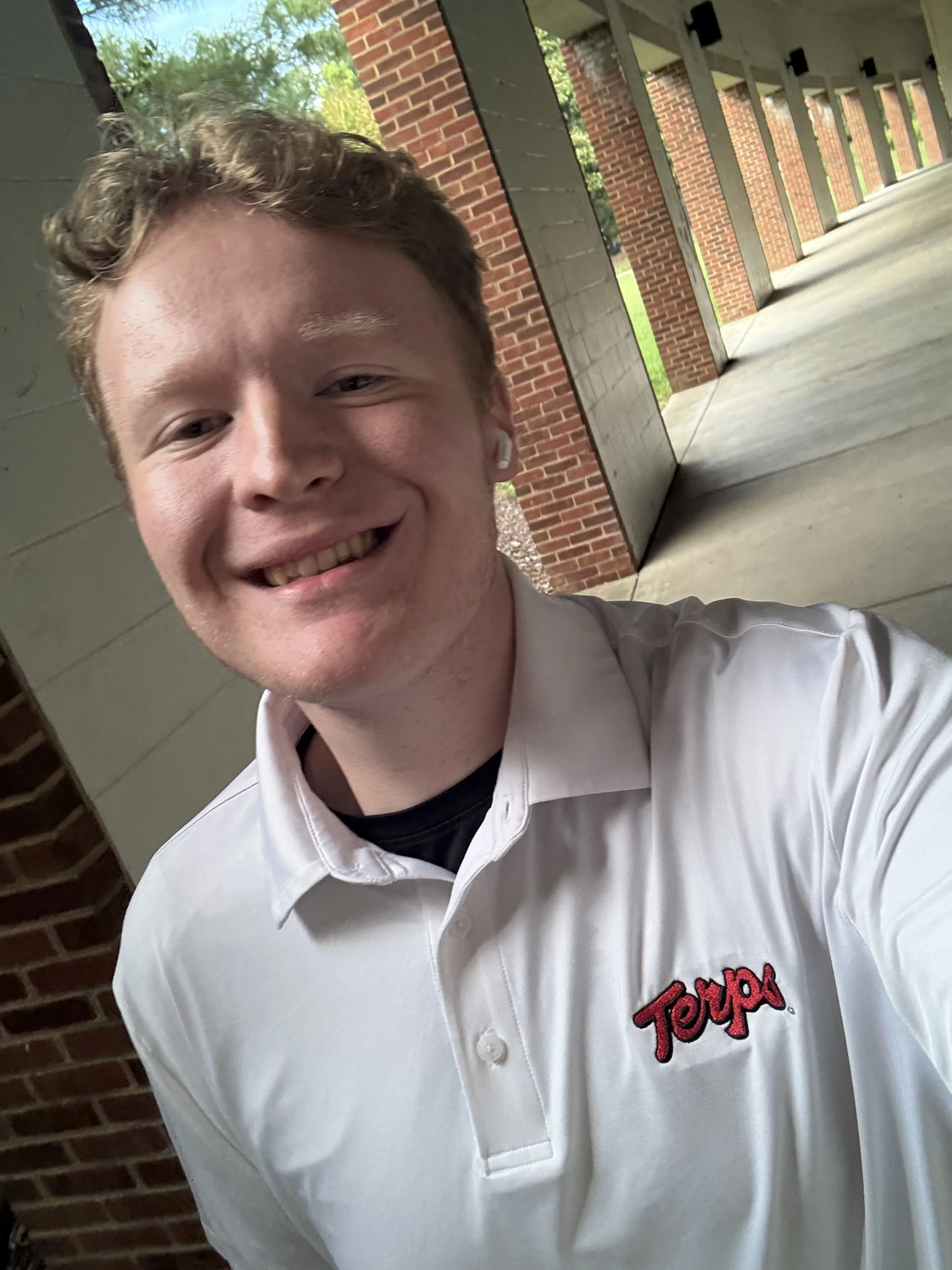 A young man with curly red hair smiling outside walking under a pathway.