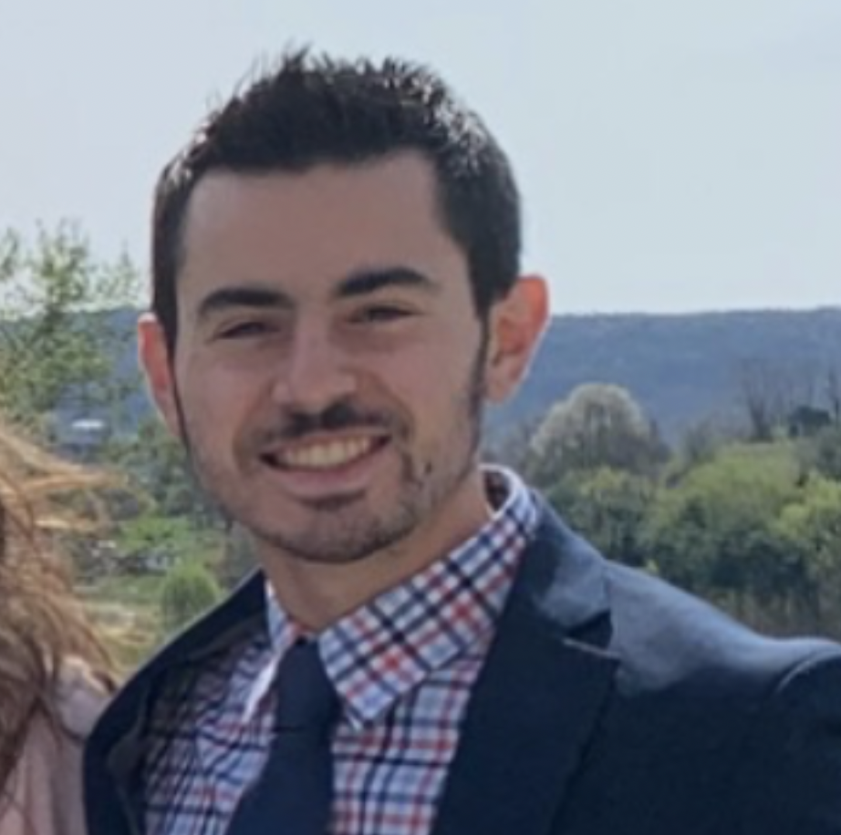 A young man with a mustache in a suit smiling with the blue sky in the backround