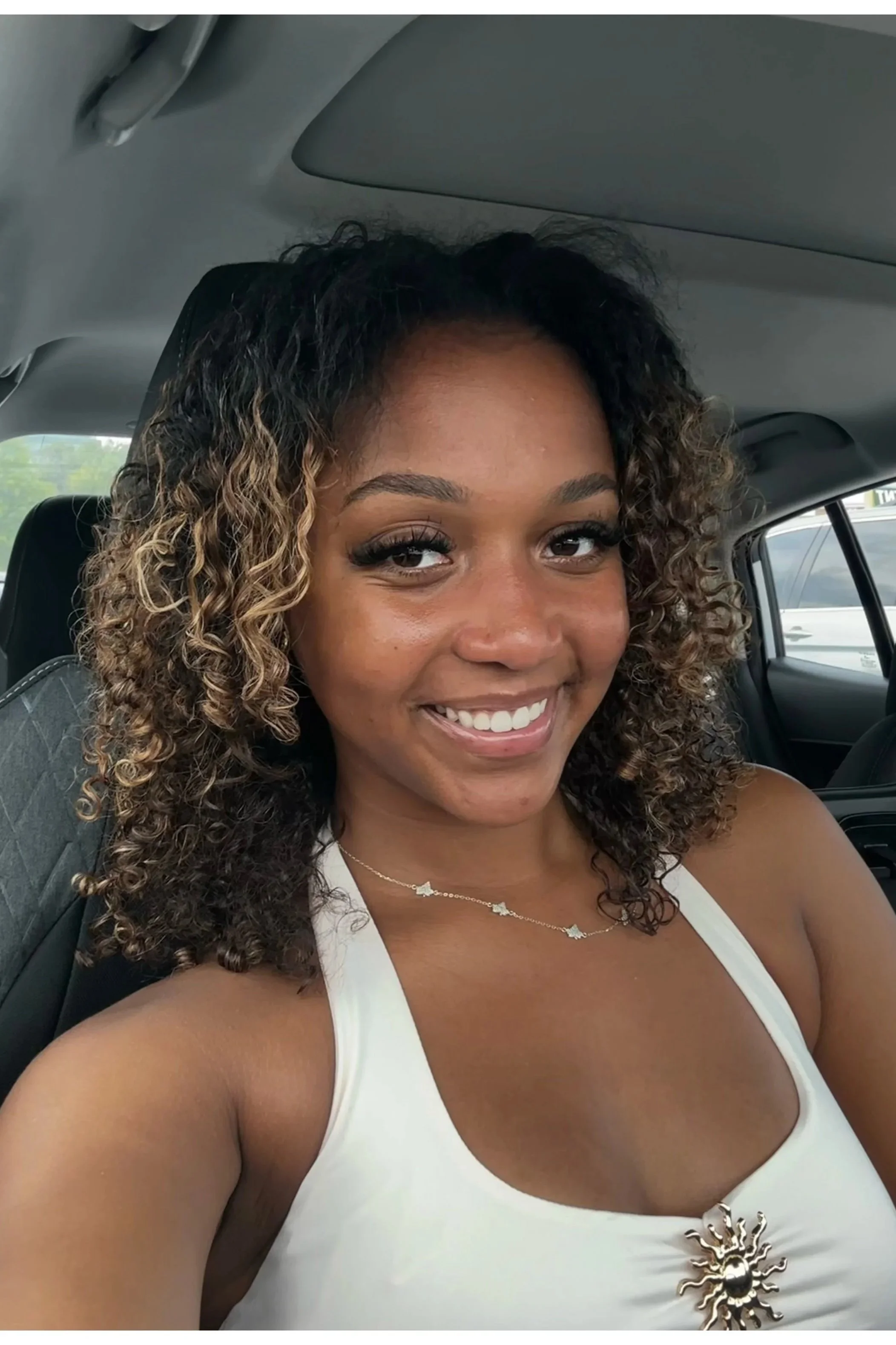 Smiling woman with curly hair and a white tank top, in a car