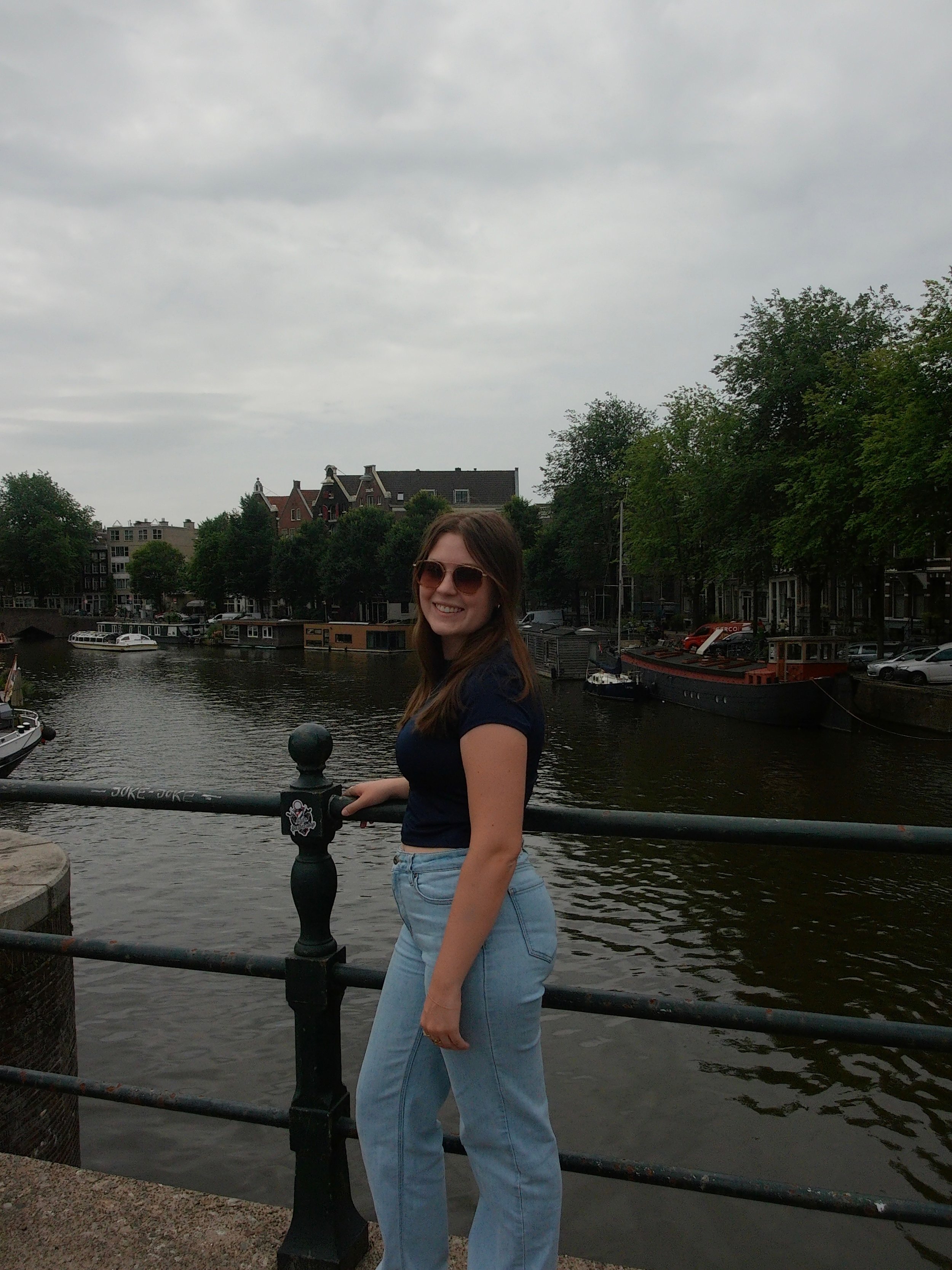 A woman wearing sunglasses, a navy blue t-shirt, and light blue jeans standing on a bridge beside a canal, with boats and trees in the background under a cloudy sky.