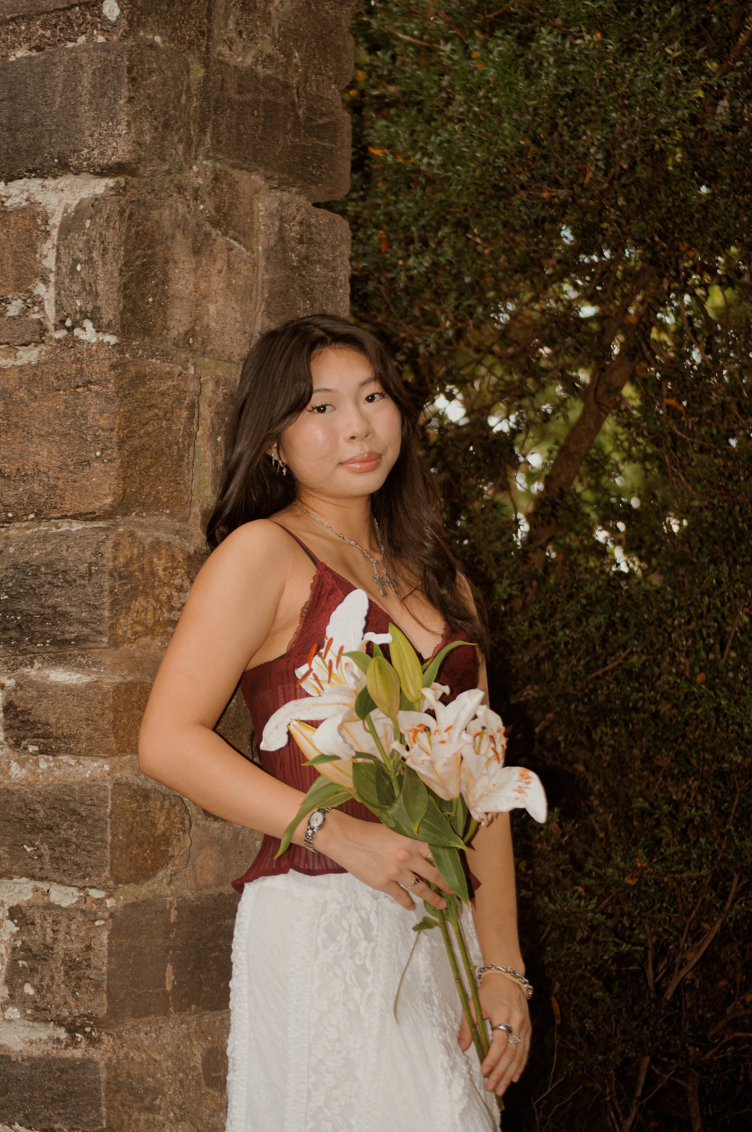 A young woman with long dark hair, wearing a white dress and holding a bouquet of flowers, smiling ourdoors