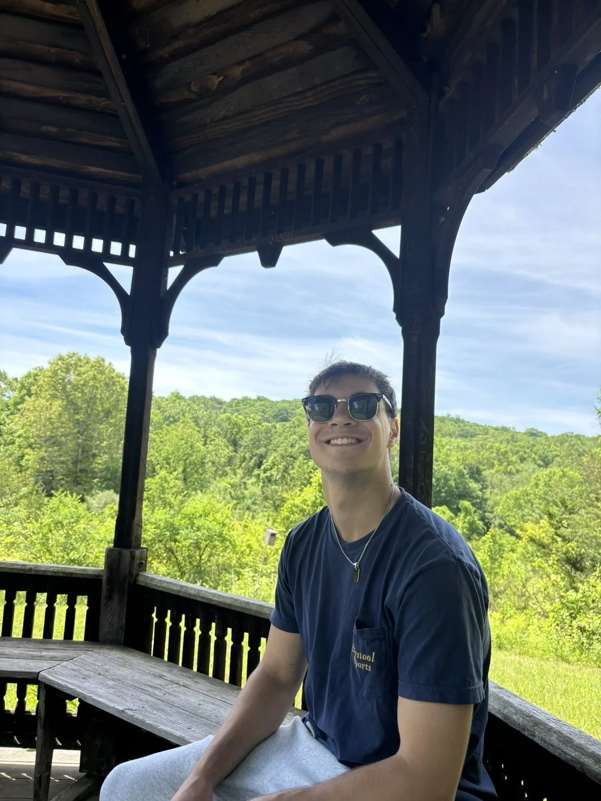 A young man sitting in a gazebo with sunglasses on and smiling