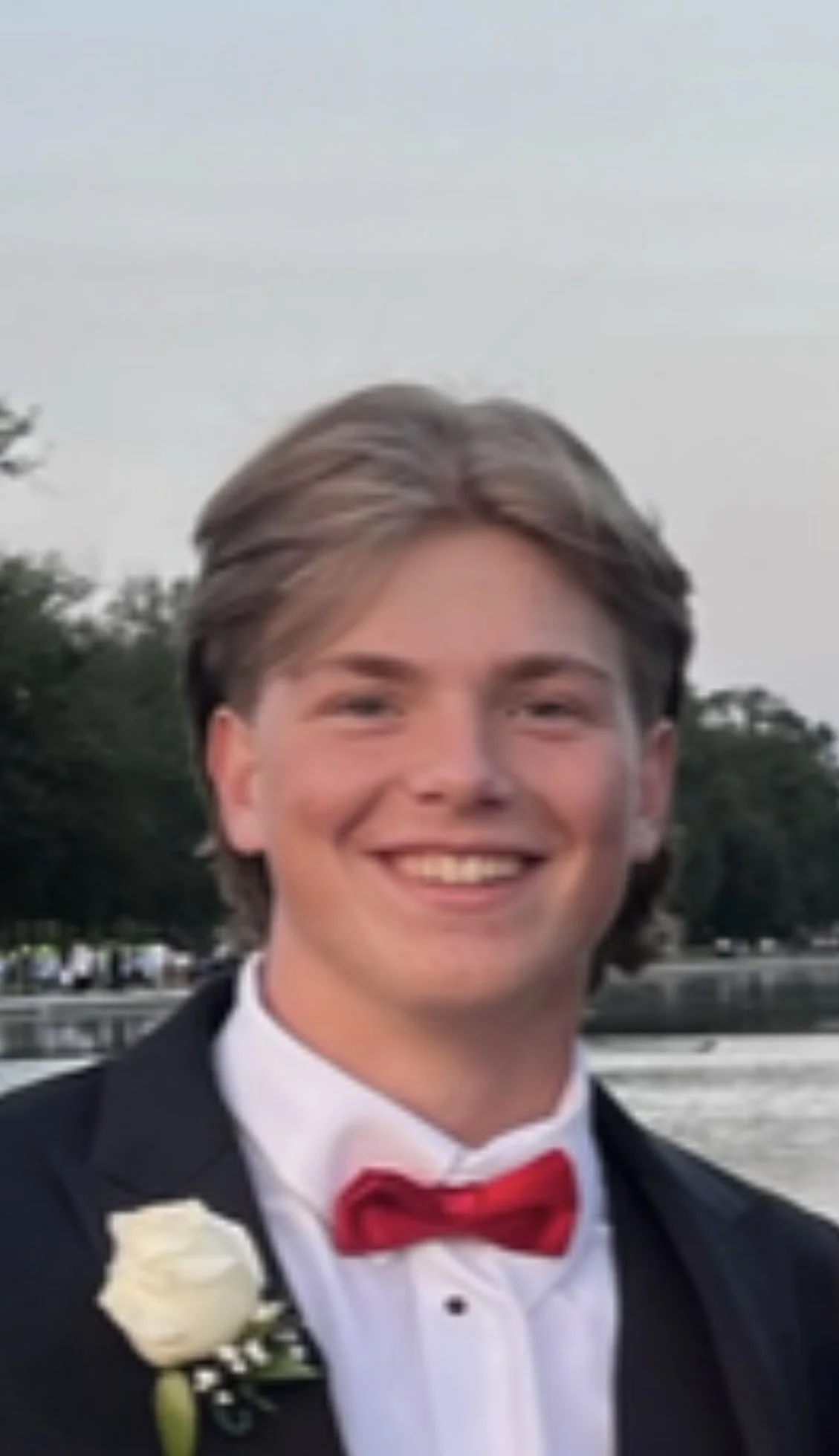 A young man with a suit on, short blond hair, and smiling with a blue sky in the backround