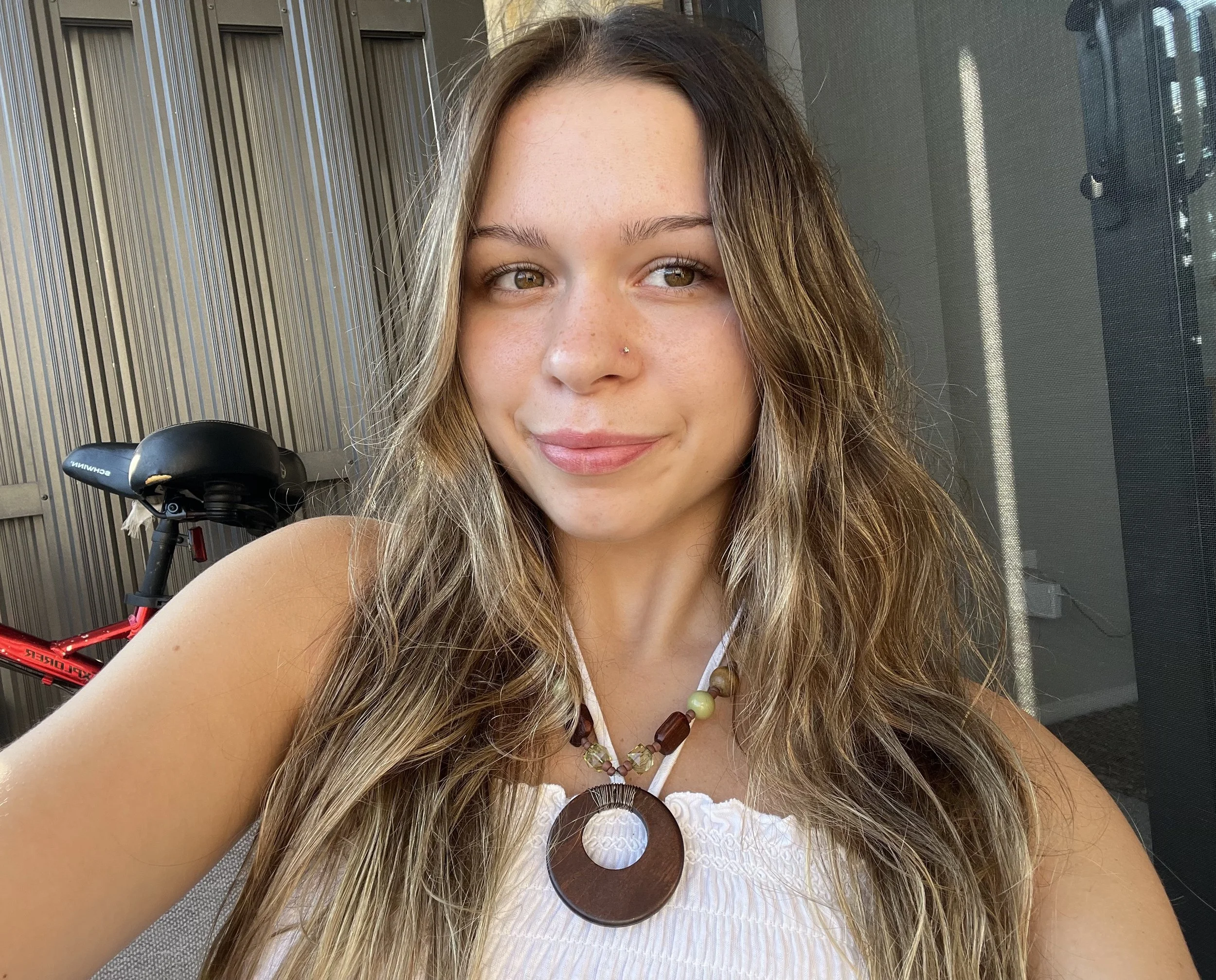 A young woman with light brown hair grinning in a selfie taken indoors, wearing a white top