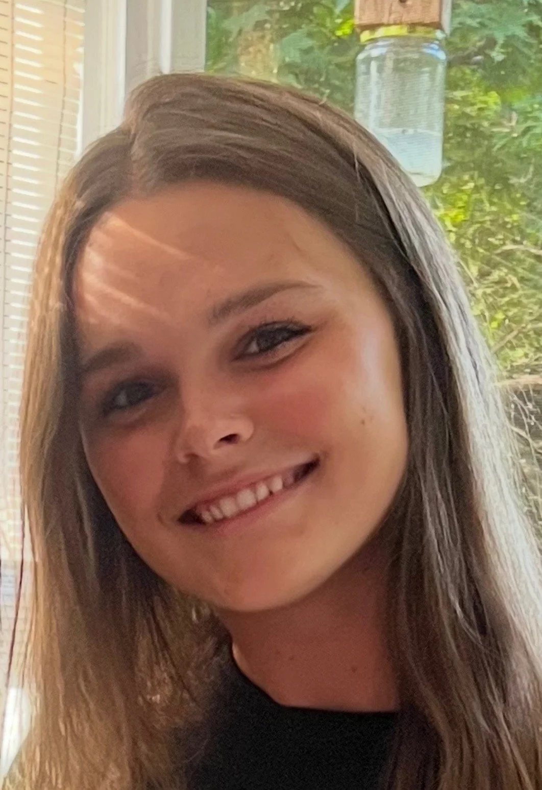 A young woman with light brown hair, smiling with greenery in the backround