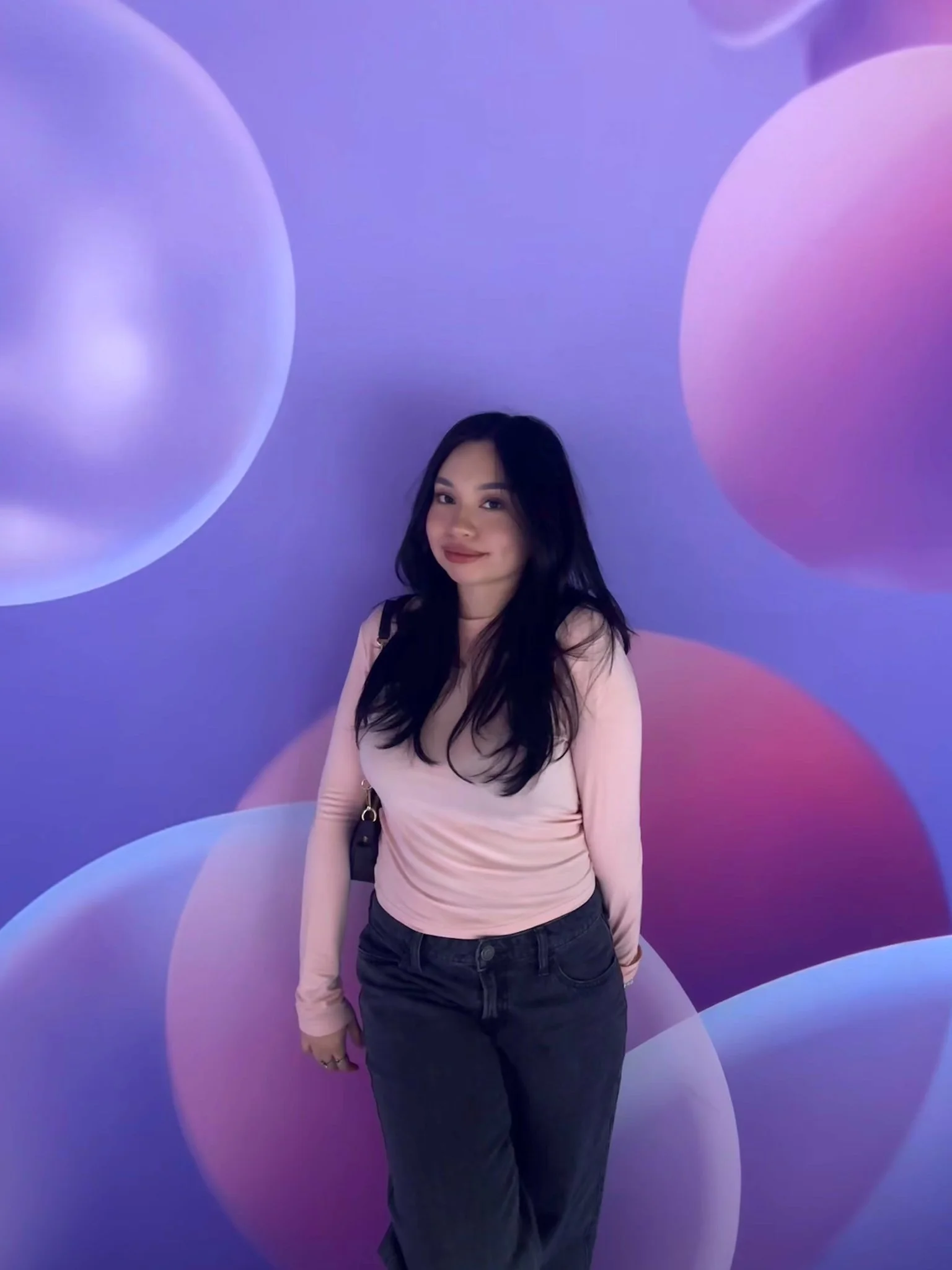 A young woman with long dark hair, smiling, surrounded by a purple and pink wallpaper indoors, standing in a white top and black pants