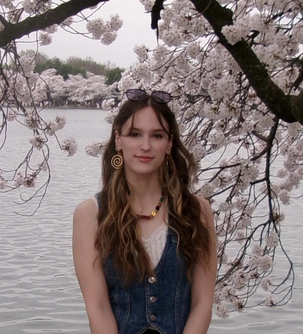 A young woman smiling with long brown hair and a black top, with cherry blossom trees in the backround