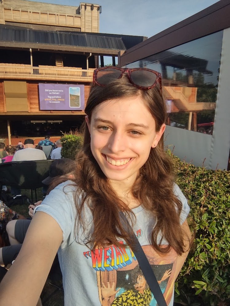 A young woman smiling outdoors with medium length brown hair and a grey shirt on