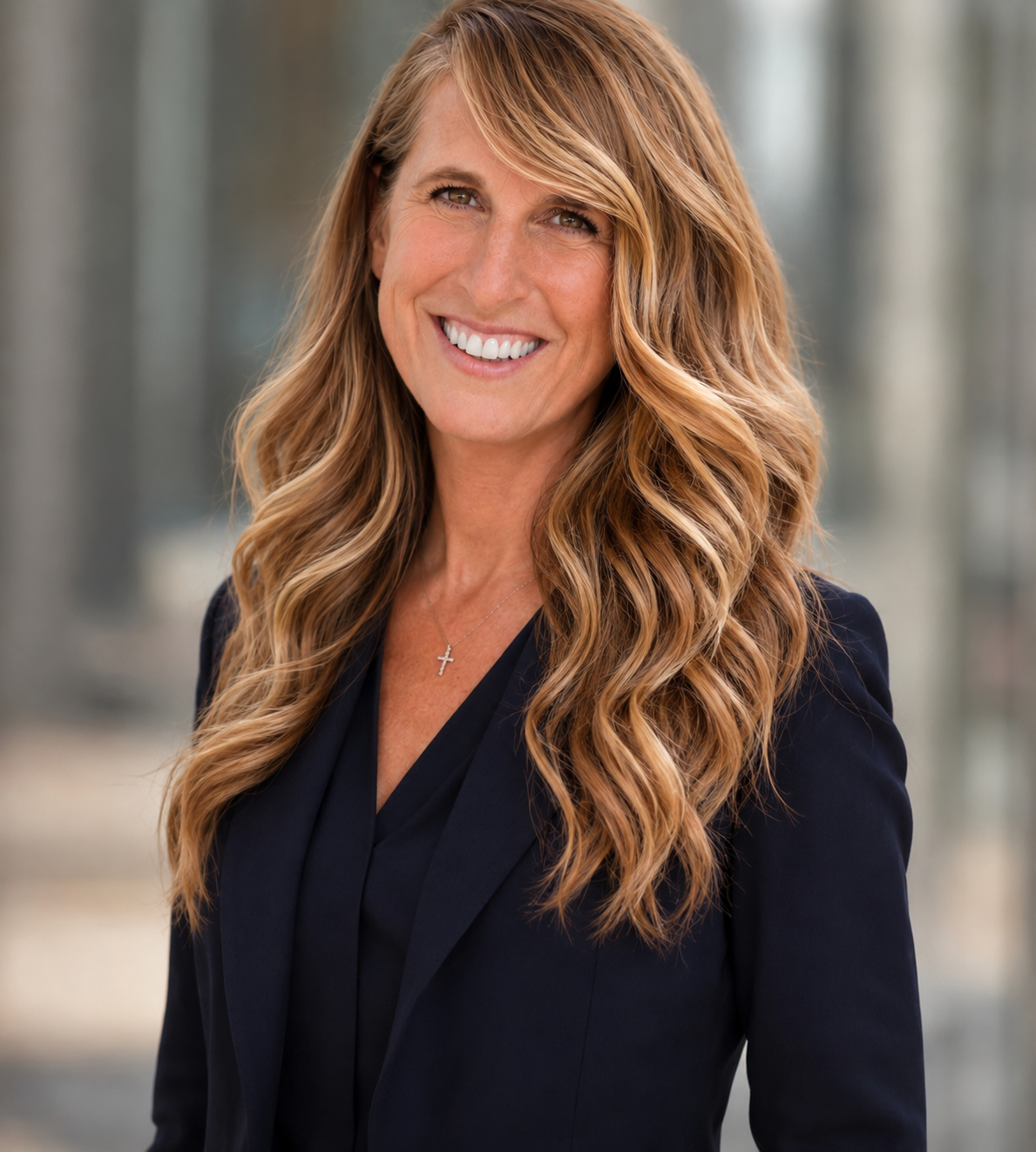 A smiling woman with long, wavy blonde hair wearing a navy blazer and a silver cross necklace, standing outdoors with a blurred background.