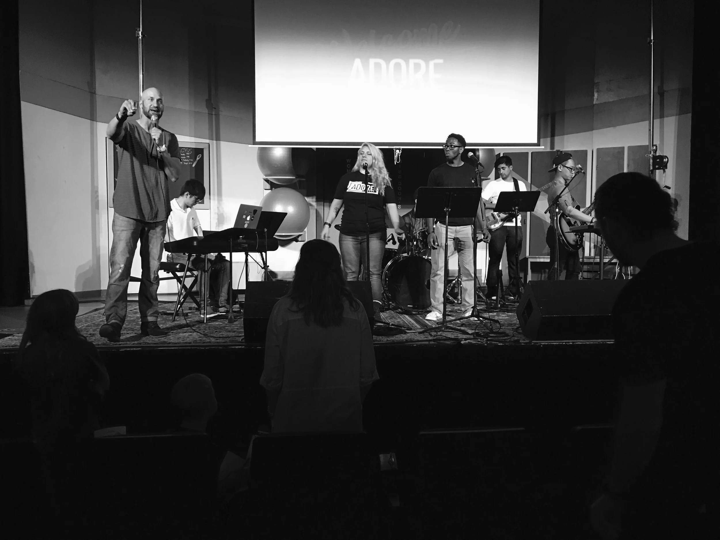 A black and white photo of a musical performance on stage with multiple musicians including a singer, guitarist, keyboardist, bassist, and drummer, with audience members in the foreground.