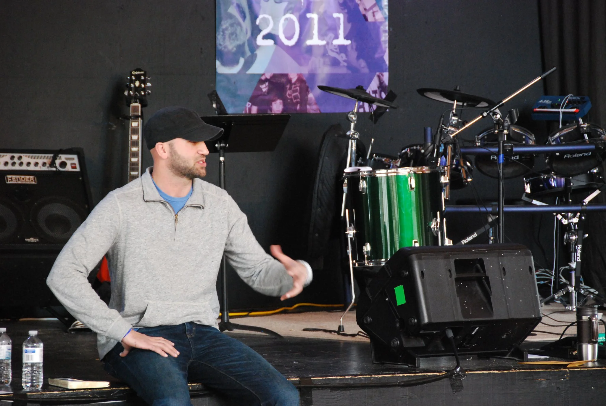 A man in a gray zip-up sweatshirt and a black cap sitting on the edge of a stage with musical instruments and equipment behind him, including a green drum set, an electric guitar, an amplifier, and electronic music gear. There is a 2011 banner hanging on the wall.