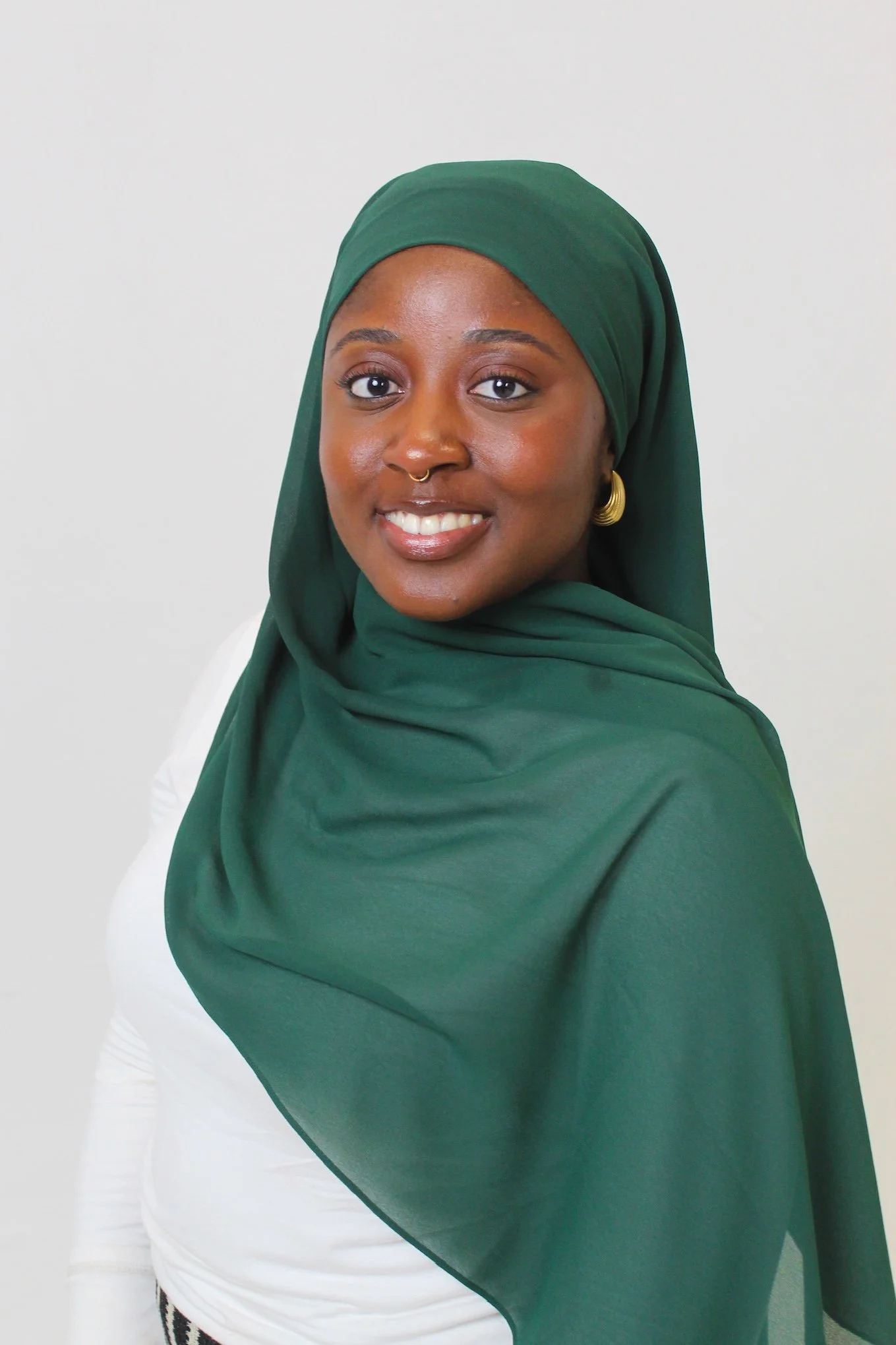 A woman wearing a green headscarf, gold hoop earrings, and a septum nose ring, smiling at the camera against a plain white background.