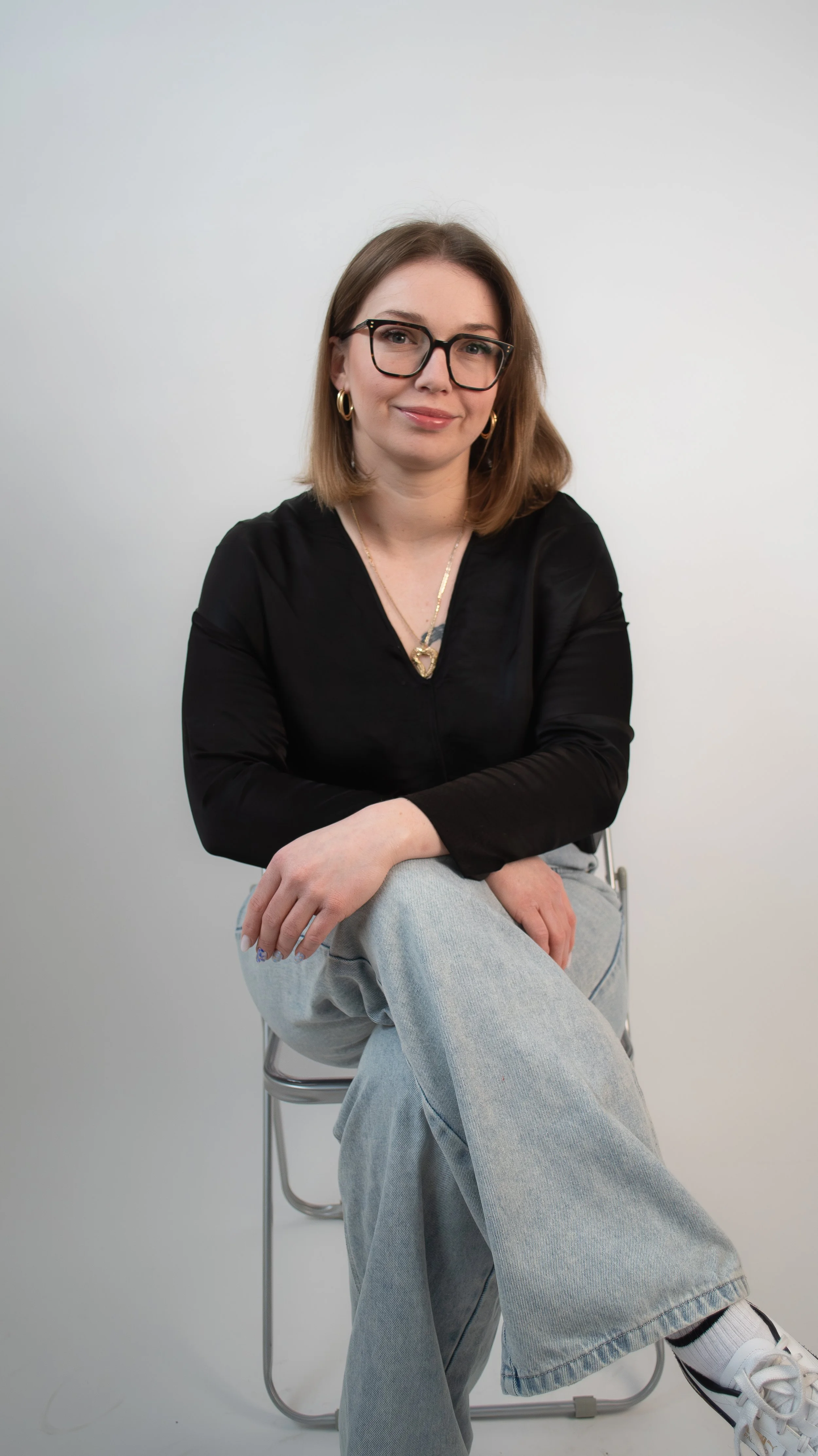 A young woman with shoulder-length brown hair, glasses, and gold hoop earrings, sitting on a silver chair against a plain white background. She is wearing a black top, light blue jeans, and white sneakers with black details, and has a relaxed, confident expression.