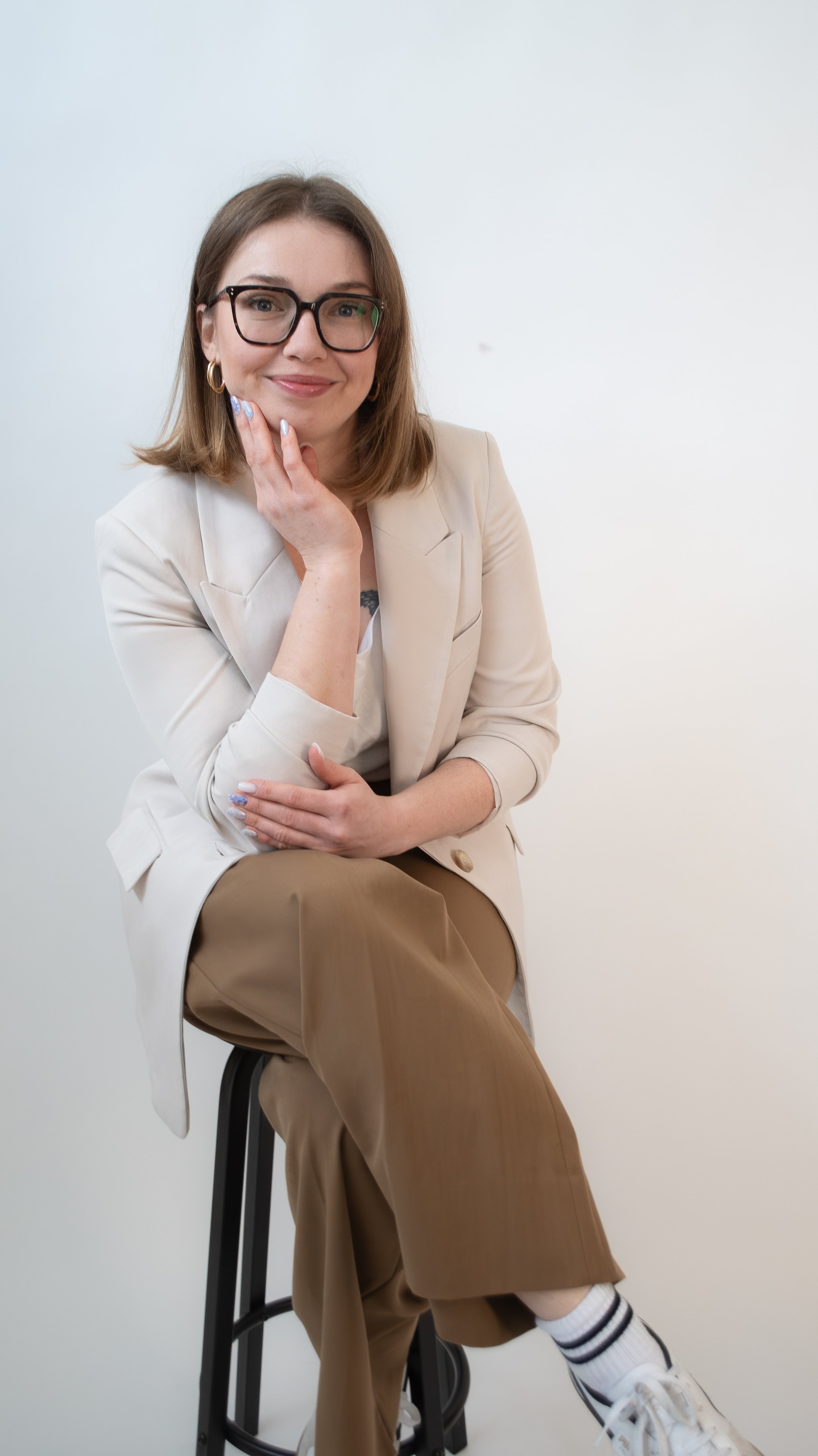 A woman with shoulder-length brown hair, wearing glasses, a beige blazer, and brown pants, sitting on a stool against a plain white background, smiling and touching her chin with her right hand.