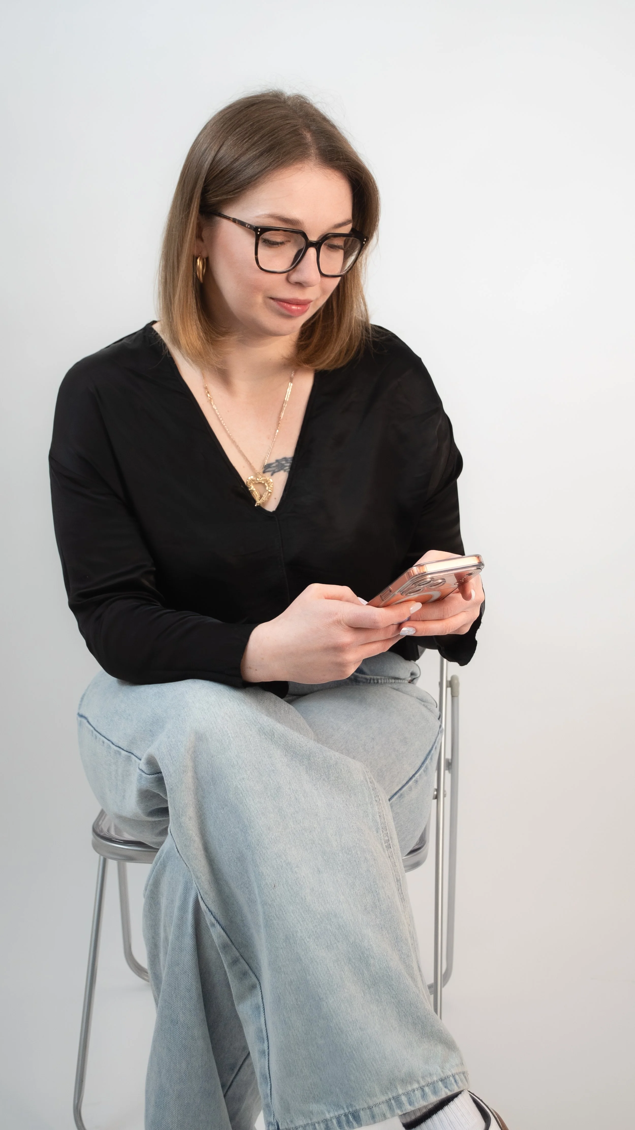 A young woman with shoulder-length brown hair, wearing glasses, a black top, and light gray jeans, is sitting on a silver chair and looking at her smartphone against a plain white background.