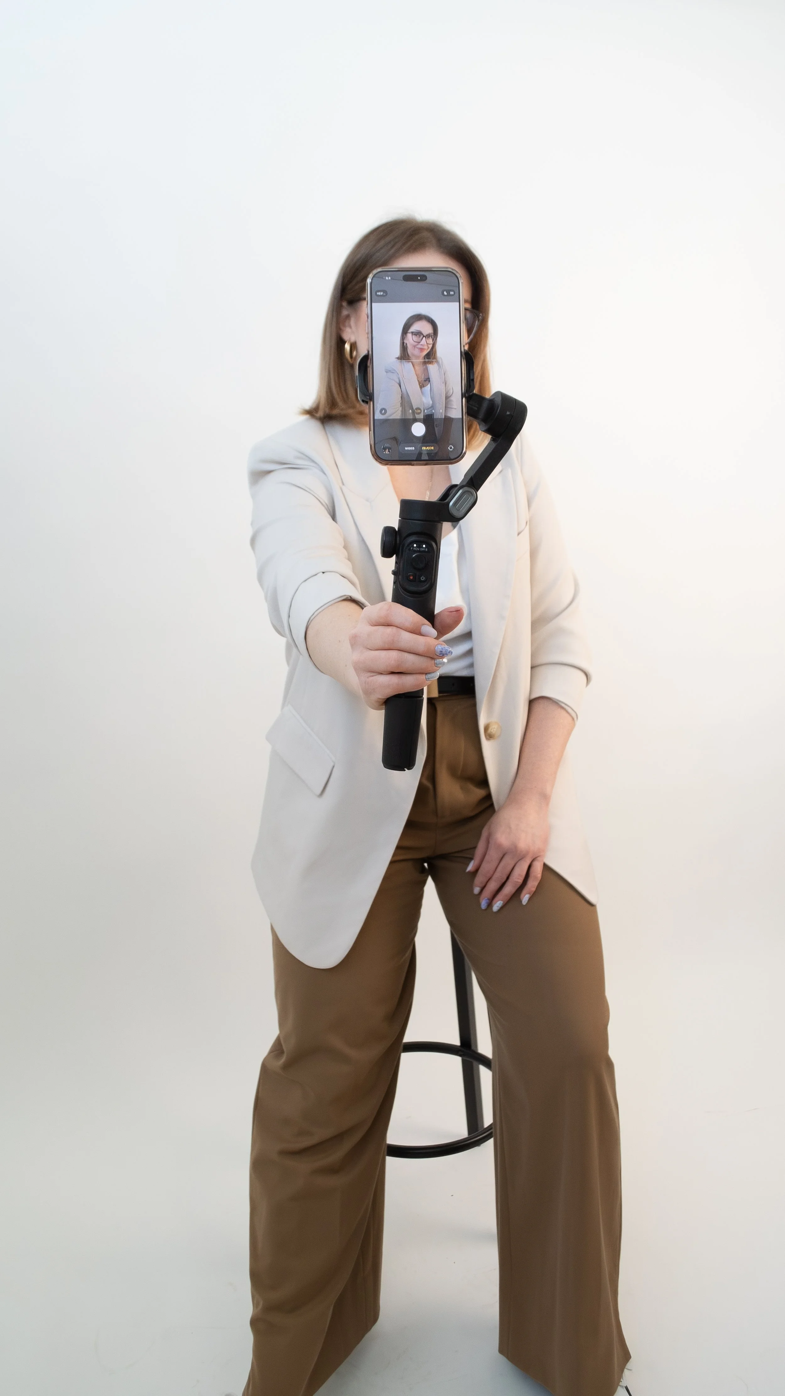 Woman in beige blazer and brown pants taking a selfie with a gimbal stabilizer and smartphone, standing against a white background.