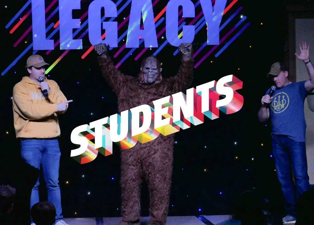 Three people on stage with a large person in a bear costume in the center, raising their fists. The person on the left is wearing a tan hoodie and cap, holding a microphone and a paper. The person on the right is wearing a blue t-shirt and cap, holding a microphone and waving. Behind them, a black backdrop features the word 'LEGACY' in large purple letters with colorful diagonal lines, and a large 3D-text that says 'STUDENTS' superimposed across the bear's torso.
