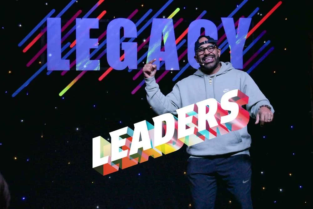 Man smiling and gesturing in front of a colorful background with the words 'LEGACY LEADERS' displayed.