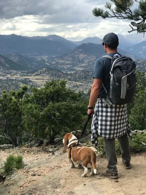 Reuben, owner of Ascent Window Tint + Clearbra in Fort Collins, Colorado, stands outdoors on a rocky trail with his Basset hound, Berkeley. They are in a mountainous area with pine trees, overlooking a valley with mountains, clouds, and a forest.