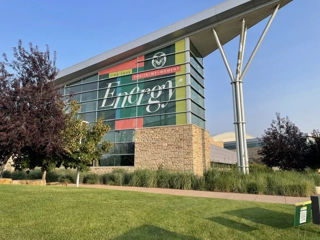 A modern, multi-story building located at the CSU Campus in Fort Collins, Colorado. Displaying a beautiful glass facade with colorful window graphics, a lush green lawn, and colorful purple leaved trees.