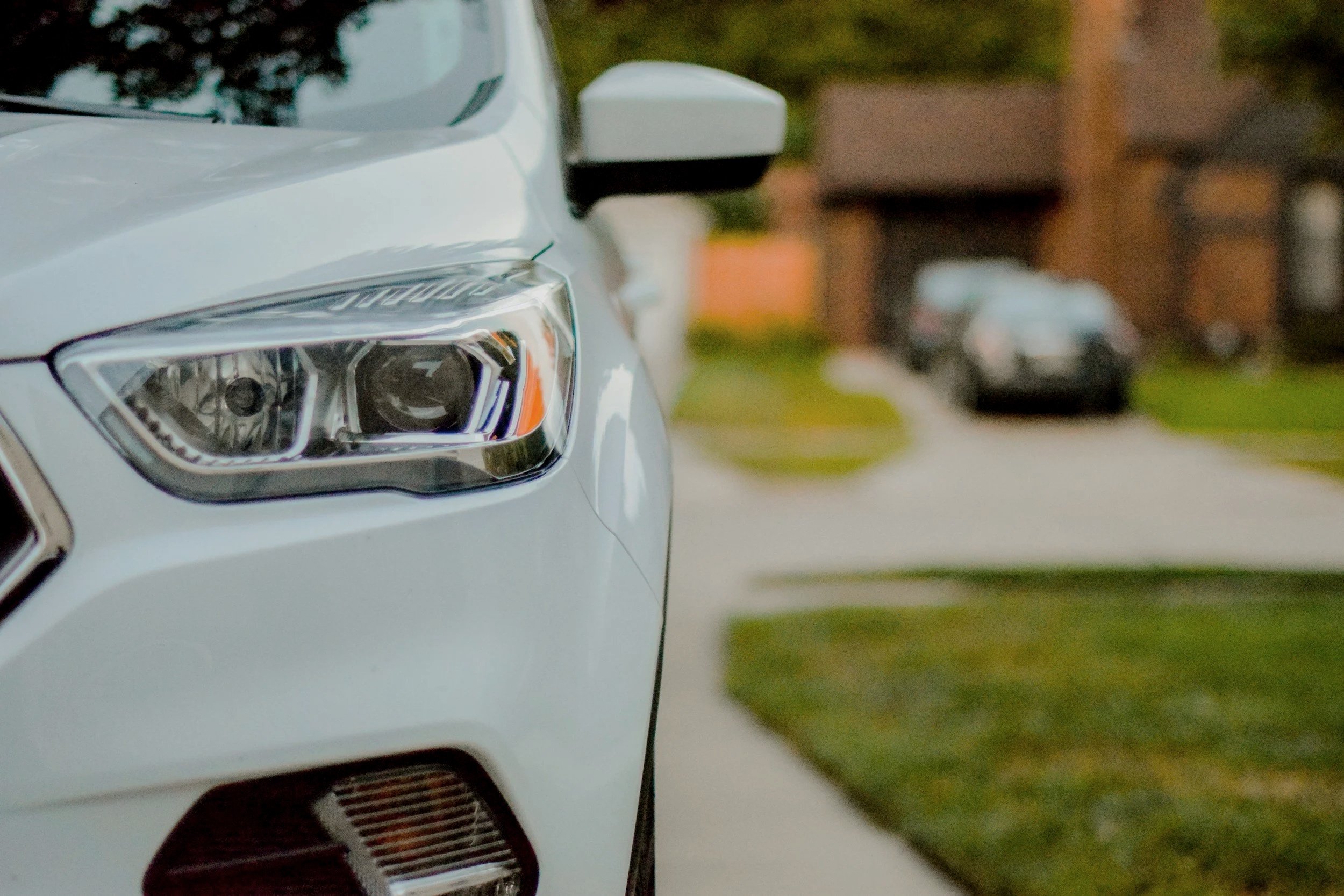 Ascent Installations offers concierge service. Close-up of the front left headlight of a white car parked on a suburban street, with other vehicles and houses blurred in the background.