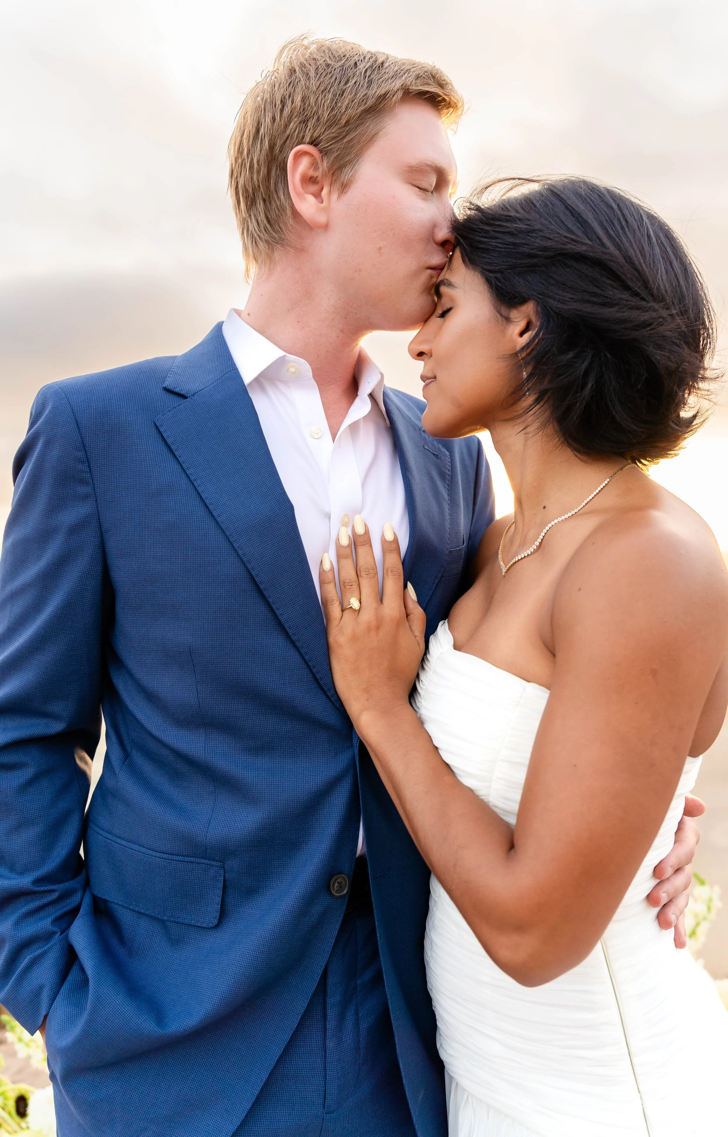 A man in a blue suit is gently kissing a woman in a strapless white dress on the forehead, with their eyes closed and a loving expression. The woman has short dark hair, and the man has light brown hair. The background is a soft, cloudy sky.