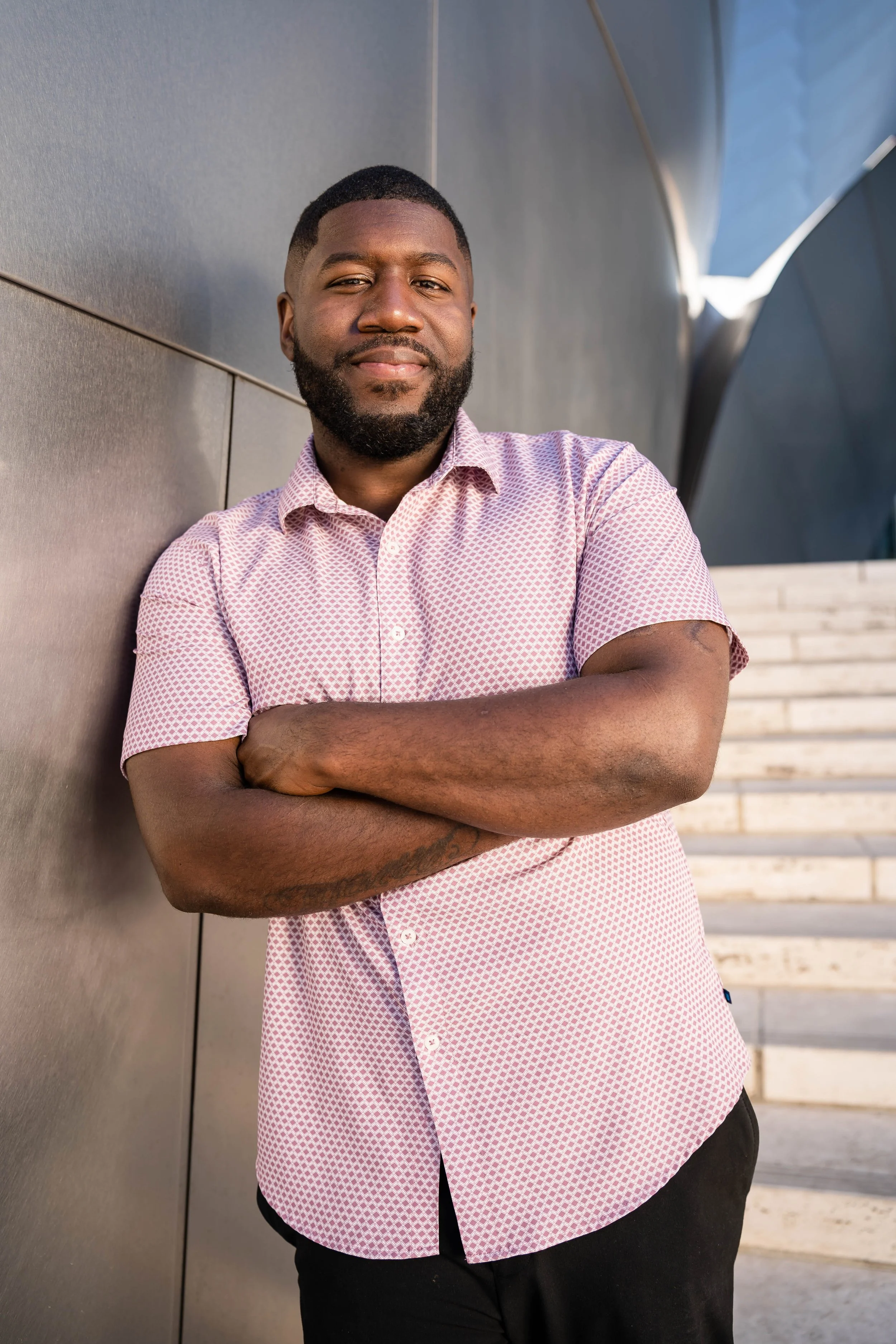 A man with a beard and short hair, wearing a pink patterned short-sleeve shirt and black pants, standing outdoors near a modern building with metallic and glass elements, smiling with his arms crossed.