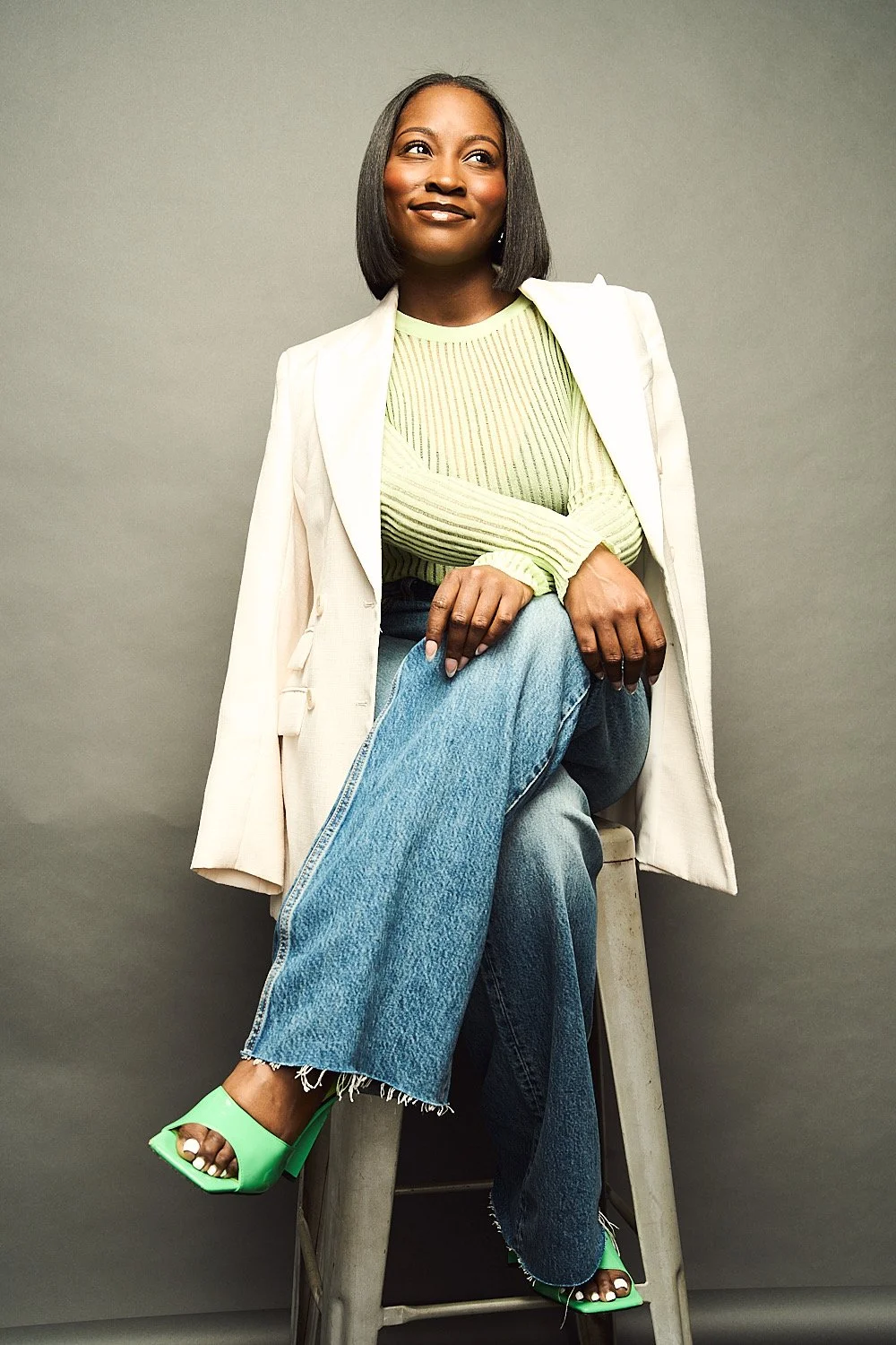 A woman with dark hair, wearing a light blazer over a green long-sleeve top, sitting on a stool against a plain gray background, with one leg crossed over the other, wearing green high-heeled sandals.