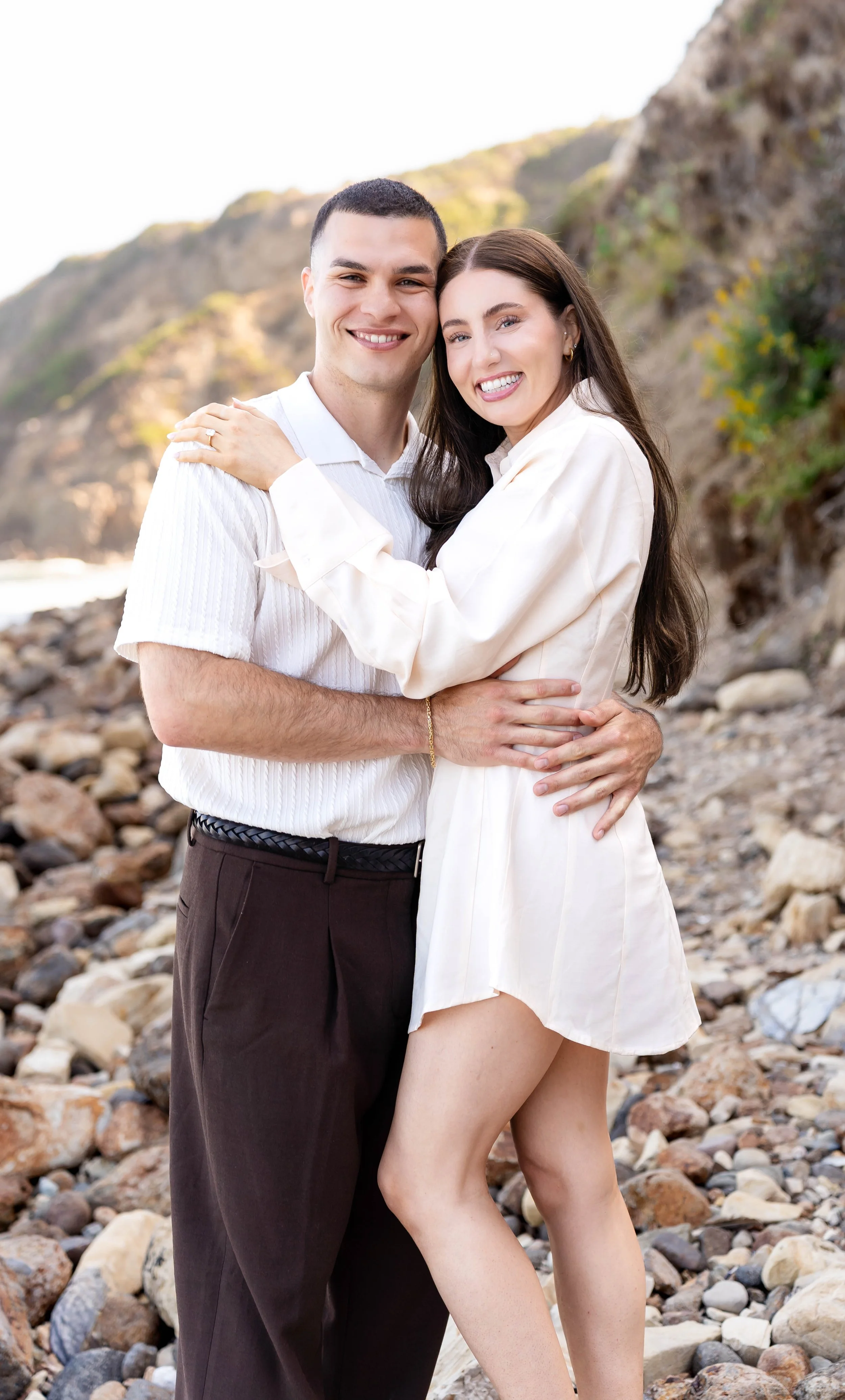 A smiling couple hugging on a rocky beach with cliffs in the background.