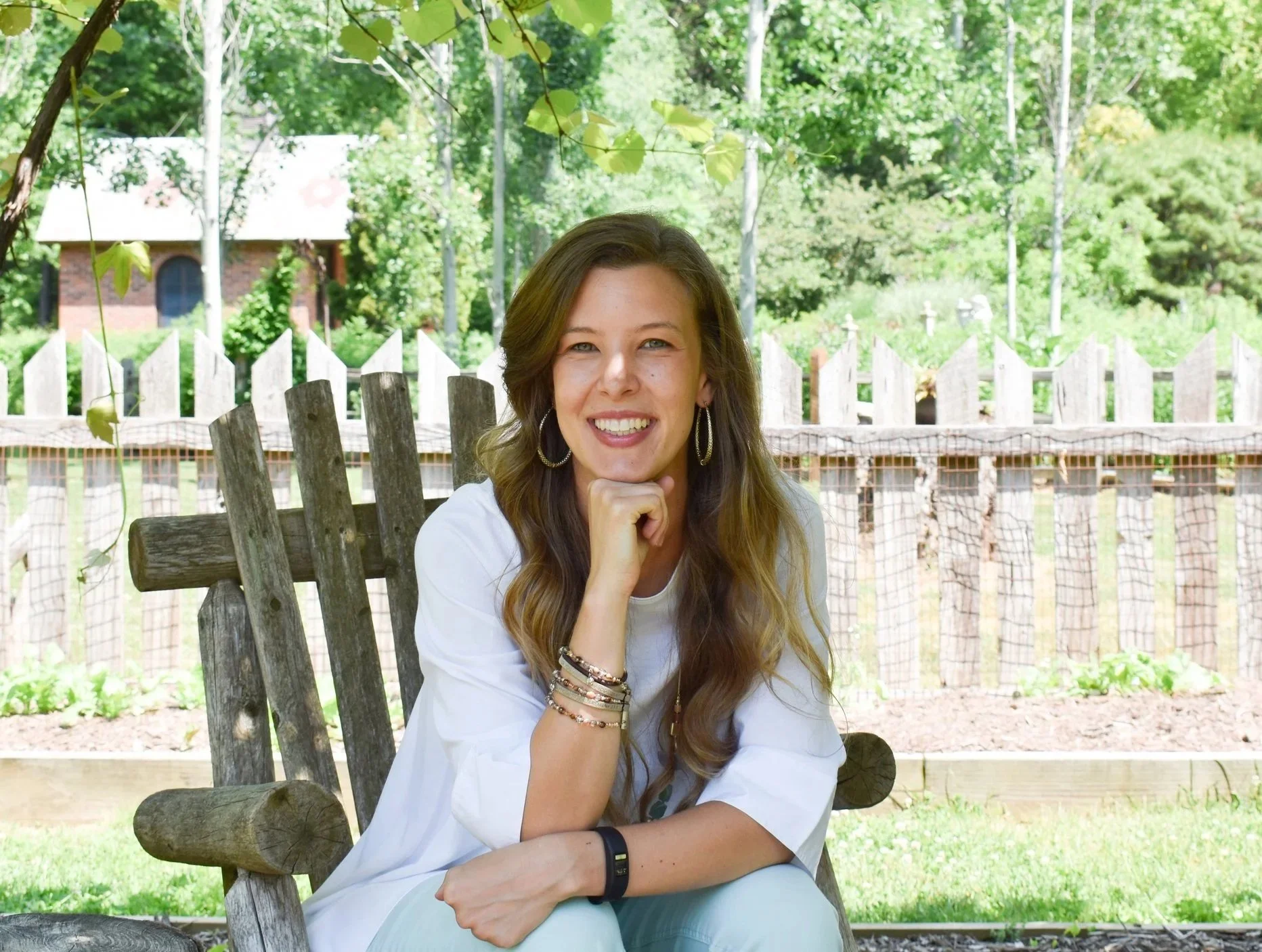 A woman sitting outdoors on a wooden bench in a backyard, smiling, with trees and a white picket fence in the background.