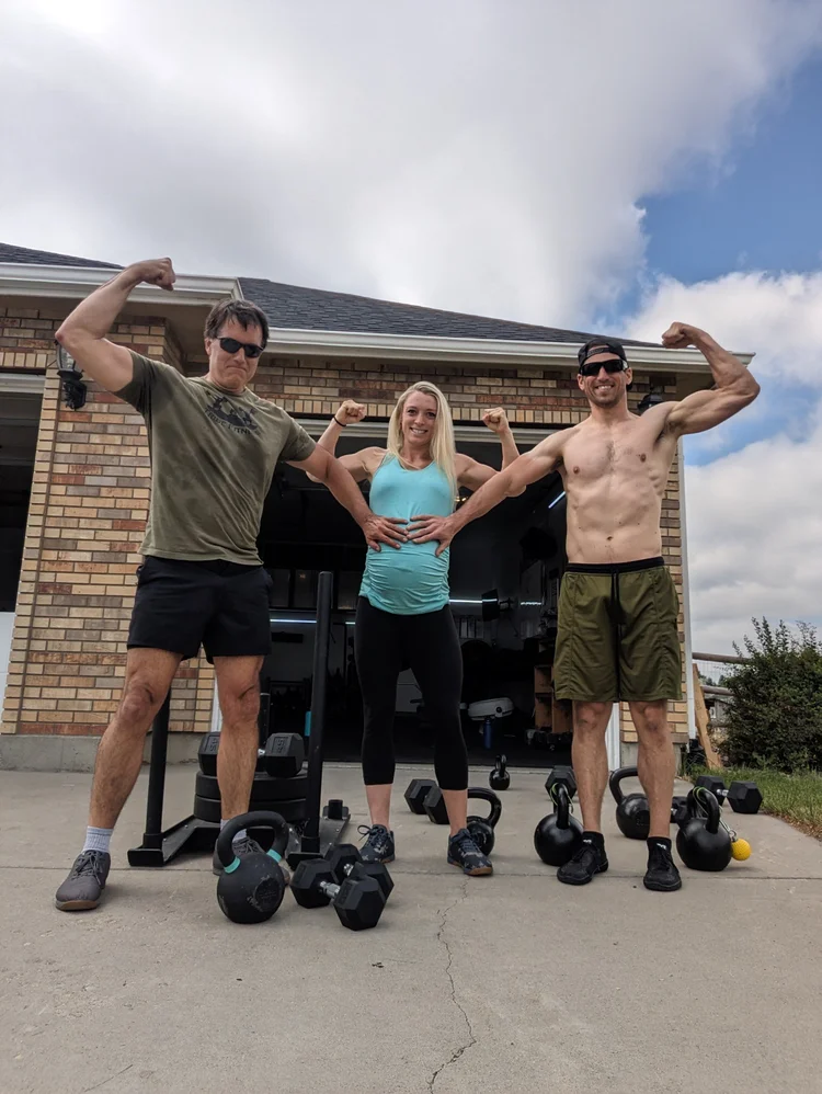 Sam Moses flexing with two training partners outside a home garage gym, with kettlebells and dumbbells on the driveway