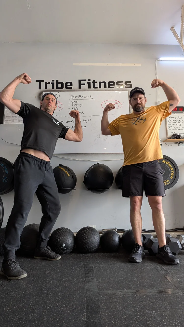 Sam Moses and a dad client flexing in front of the Tribe Fitness whiteboard