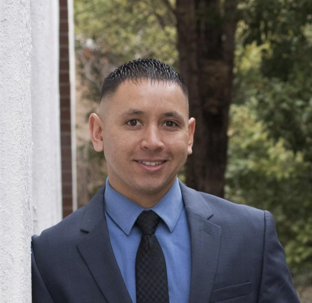 Isaiah Vargas Gunbuddy CEO in a dark suit, blue shirt, and black tie standing outdoors with trees in the background, smiling at the camera.