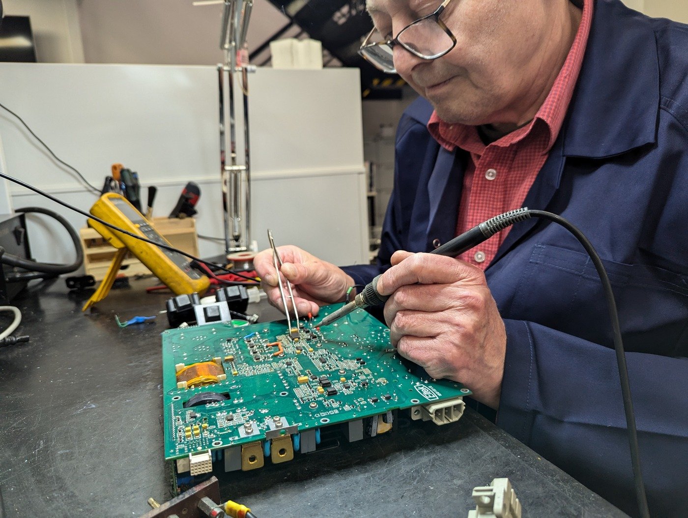 A man wearing glasses and a blue lab coat soldering electronic components on a green circuit board at a workbench, with various tools and equipment in the background.
