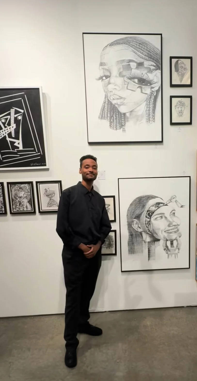 ngelo Vazquez stands in a gallery wearing black, posing in front of a wall of framed black-and-white drawings, including two large surreal portrait pieces.