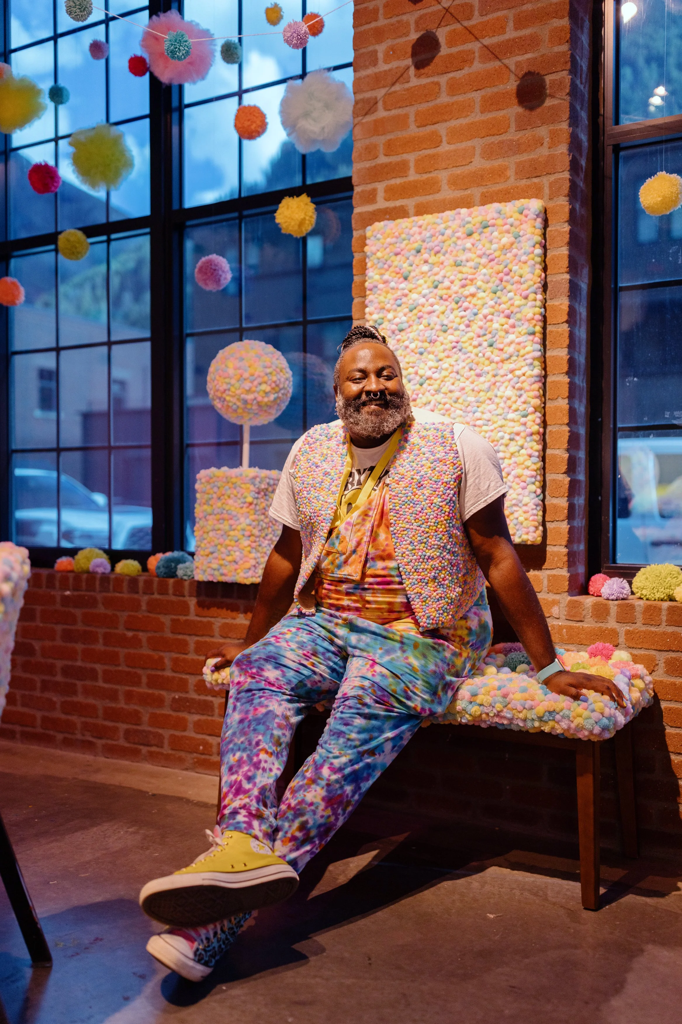 Mister Larrie sits smiling on a bench covered in soft, multicolored pom-pom textures inside a gallery space.